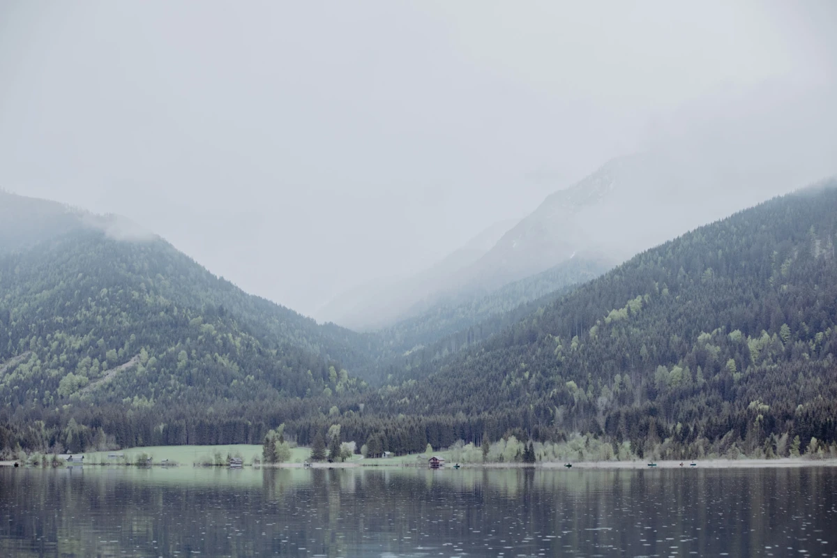 Bergsee in nebliger Waldlandschaft; ruhiges Wasser spiegelt Wälder und bergige Kulisse wider.