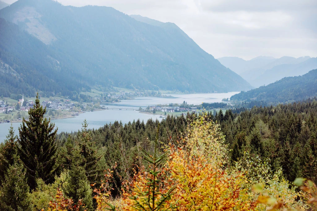 Gebirgslandschaft mit See und Dorf am Ufer, im Vordergrund bunte Herbstwälder und dunkle Tannen.