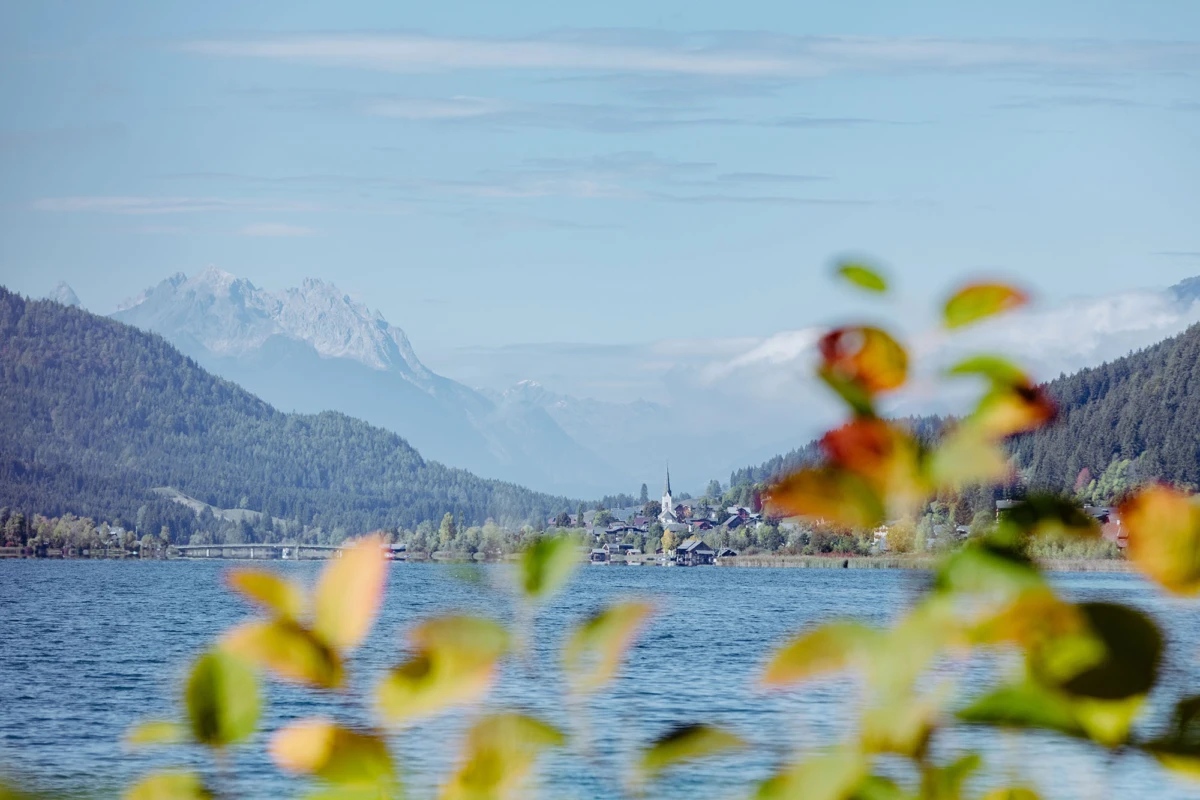 See mit Dorf am Ufer, Bergketten im Hintergrund, schneebedeckte Gipfel; Vordergrund unscharfe Herbstblätter.