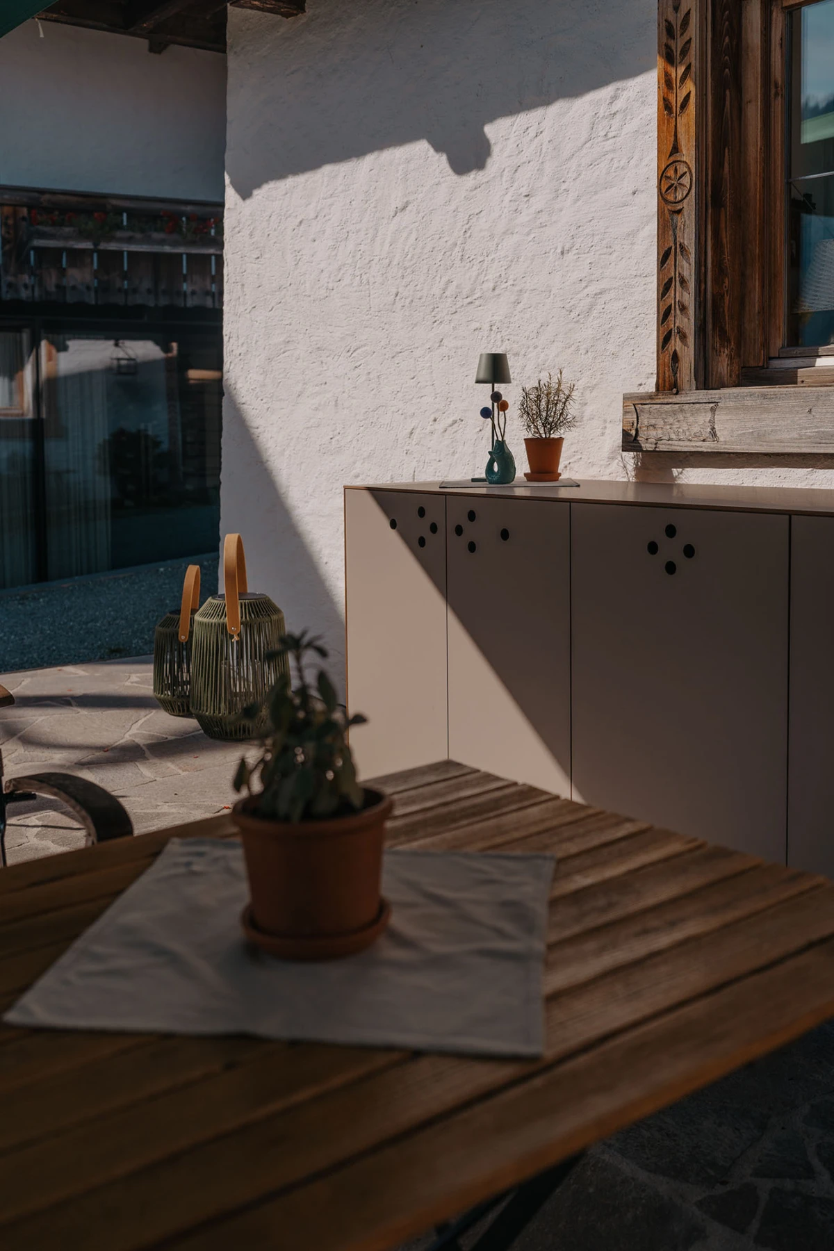 Terrasse: Holztisch mit Topfpflanze auf Serviette; beige Schrankfront; Wand mit geschnitztem Fensterrahmen; warmes Sonnenlicht.