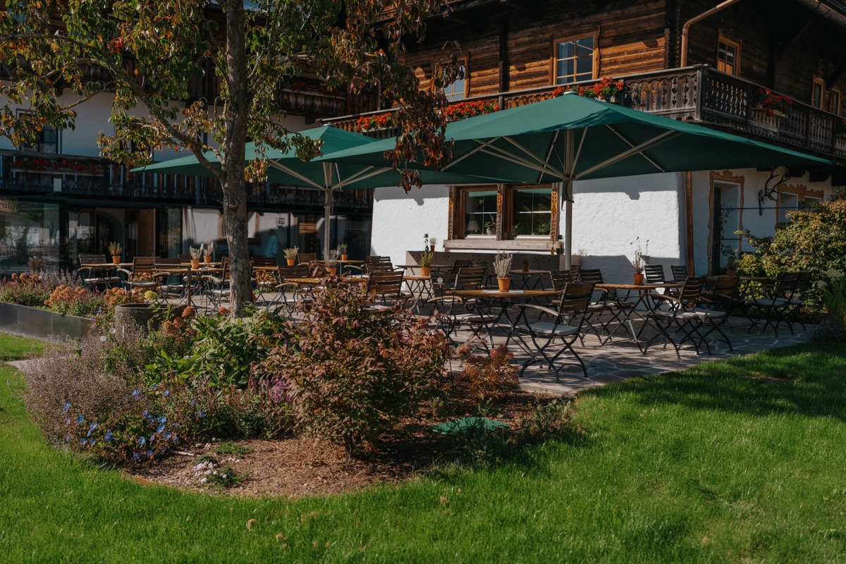 Außenterrasse eines Alpenstil-Gasthauses mit leeren Holztischen unter grünen Sonnenschirmen; Garten im Vordergrund.