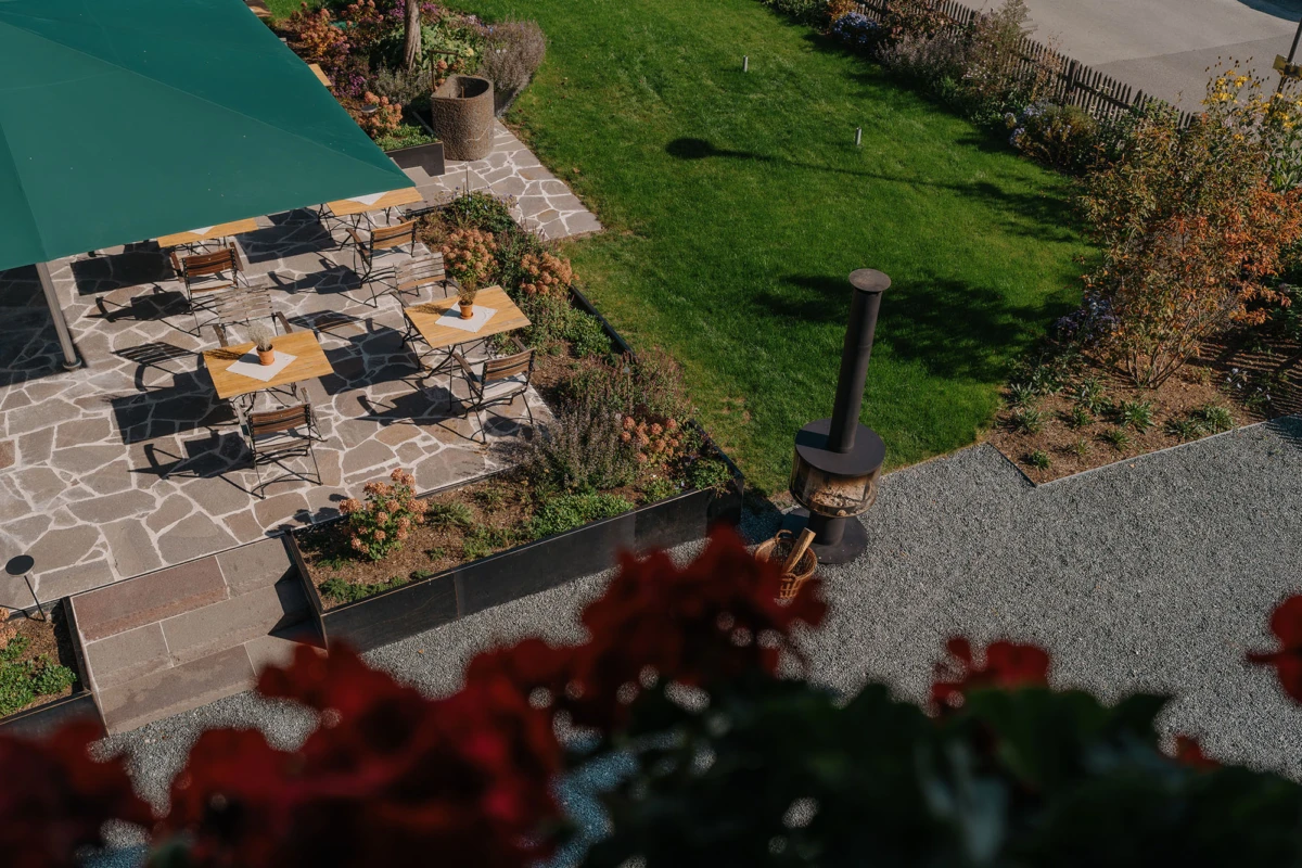 Gartenterrasse aus Stein mit vier Holztischen und Stühlen unter grünem Sonnenschirm; Feuerstelle auf Kies, grüne Rasenfläche im Hintergrund.