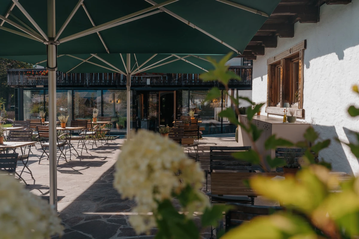 Außen-Caféterrasse mit Holztischen und Stühlen, großen grünen Sonnenschirmen, Steinboden, weißem Gebäude und blühenden Pflanzen im Vordergrund.
