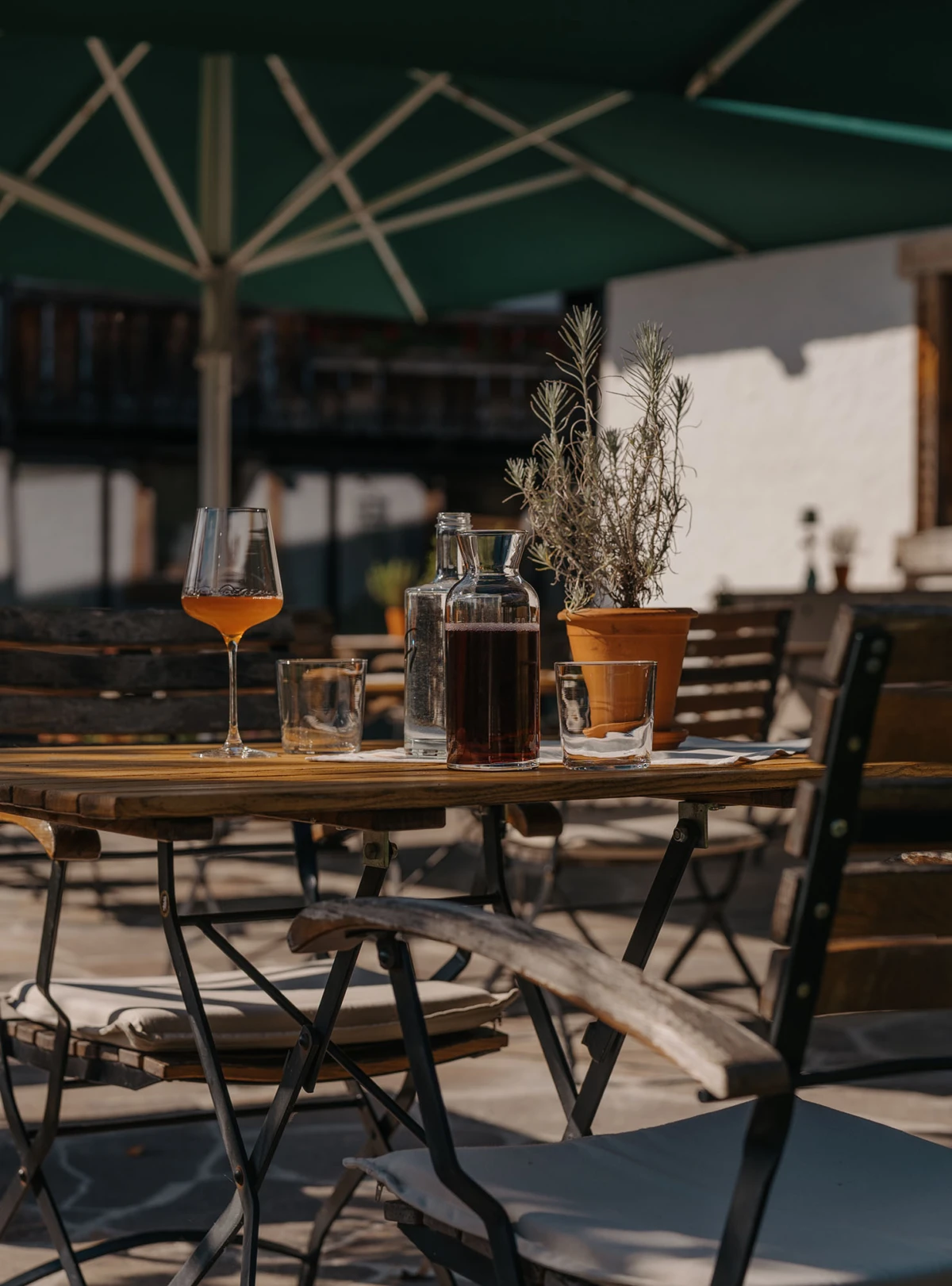 Holztisch im Außenbereich eines Cafés mit Weinkaraffe, Gläser und Rosmarinpflanze unter grünem Sonnenschirm