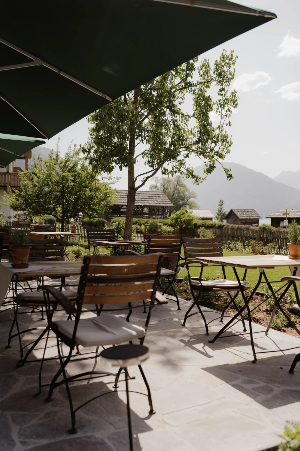 Außenterrasse eines Landcafés mit Holzstühlen und Metalltischen unter großen Sonnenschirmen; grüne Bäume und Berglandschaft im Hintergrund.
