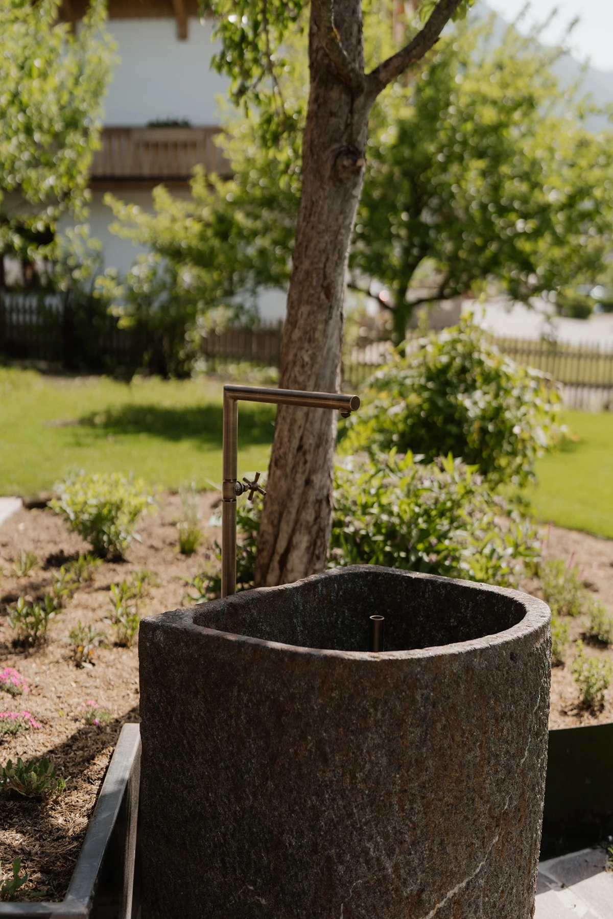 Runder Steintrog mit Metall-Handpumpe im Garten, Bäume im Hintergrund.