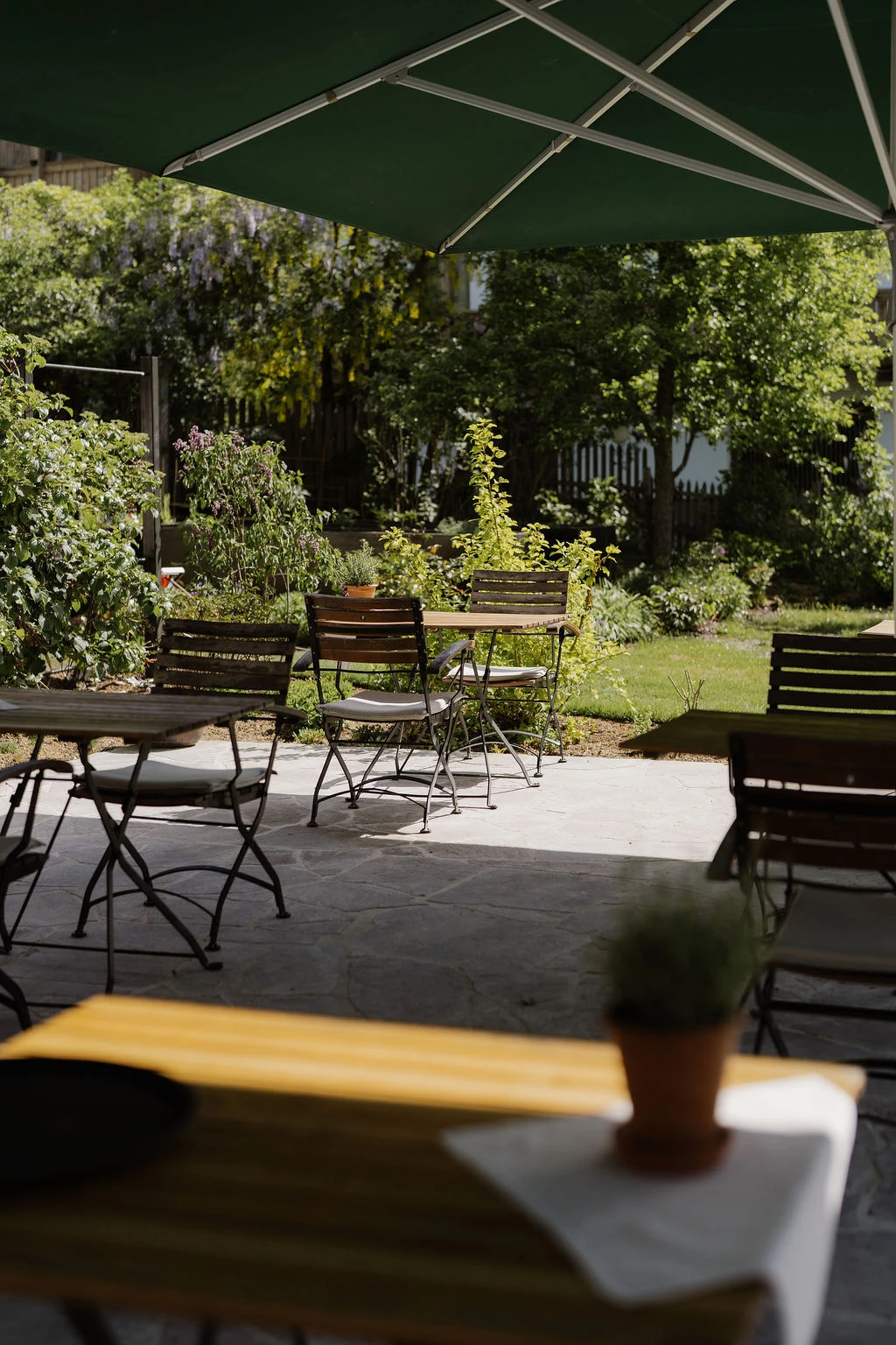 Leerer Außen-Terrassenbereich mit Holztischen und Klappstühlen unter grünem Sonnenschirm, umgeben von Gartenpflanzen.