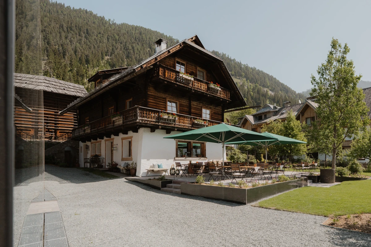 Alpenchalet aus Holz mit Terrasse und grünem Sonnenschirm; Gartenrestaurant vor Berglandschaft.