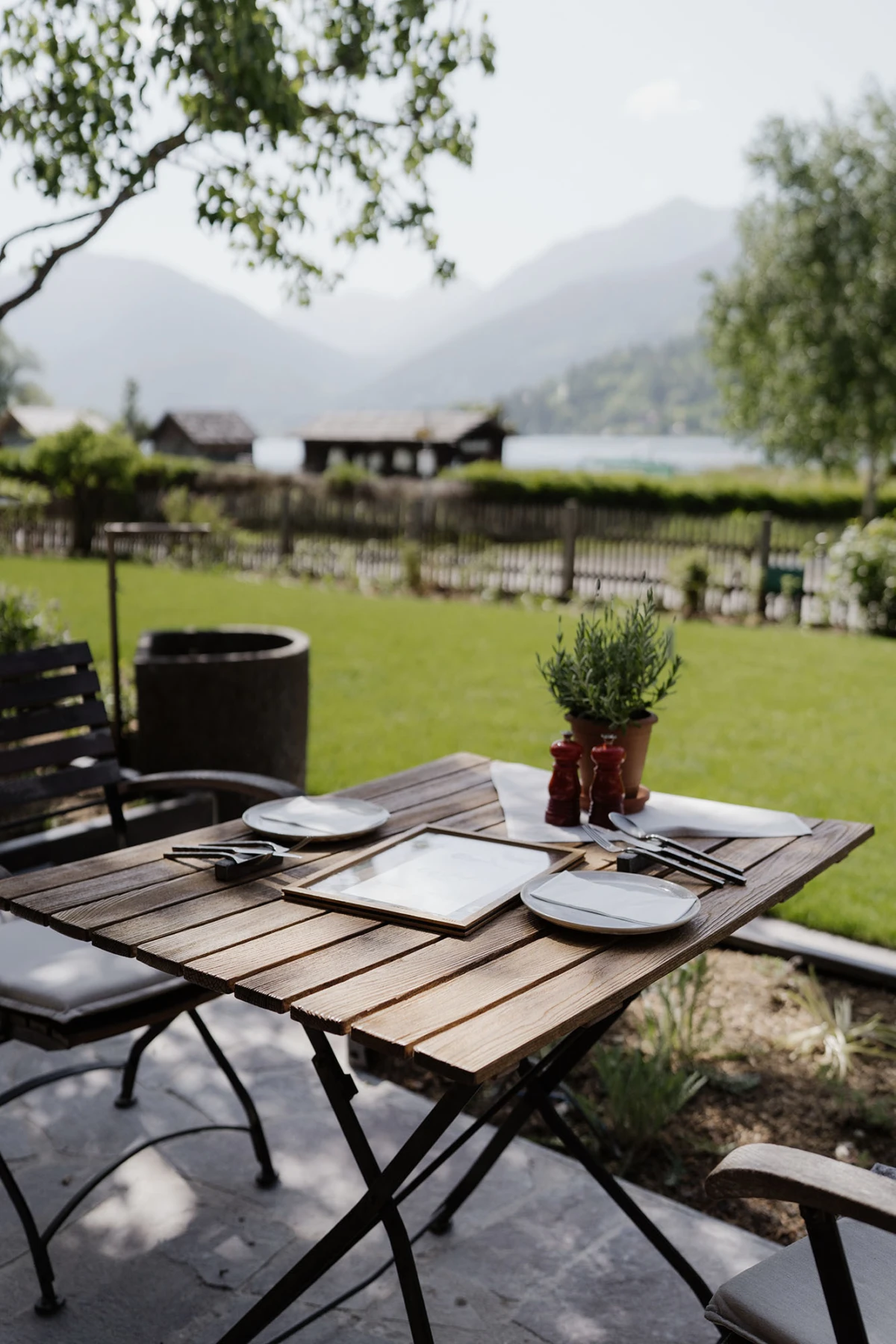 Gedeckter Holztisch im Freien mit Tellern und Besteck, Pfeffer- und Salzstreuer, Topfpflanze; Gartenblick auf See und Berge.
