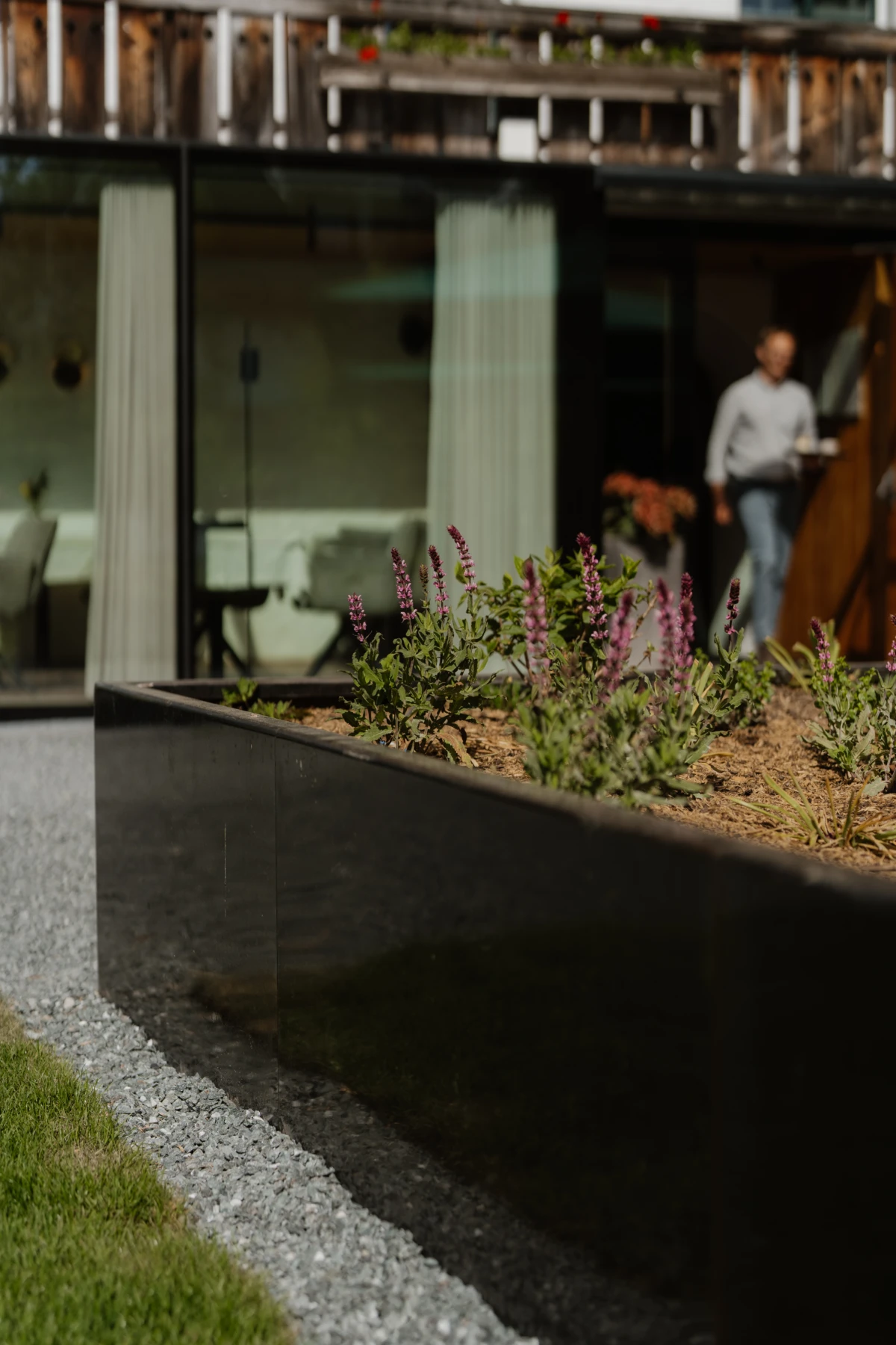 Schwarzes Metallpflanzbeet mit Lavendel im Vordergrund vor modernem Haus; im Hintergrund steht eine Person am Eingang.