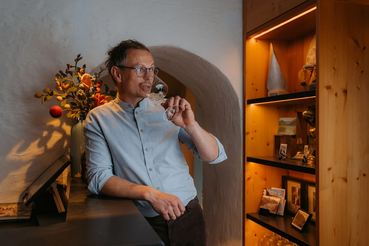 Person mit Brille trinkt Wein aus Weinglas an dunkler Theke, neben Vase mit Trockenblumen und Holzregal.