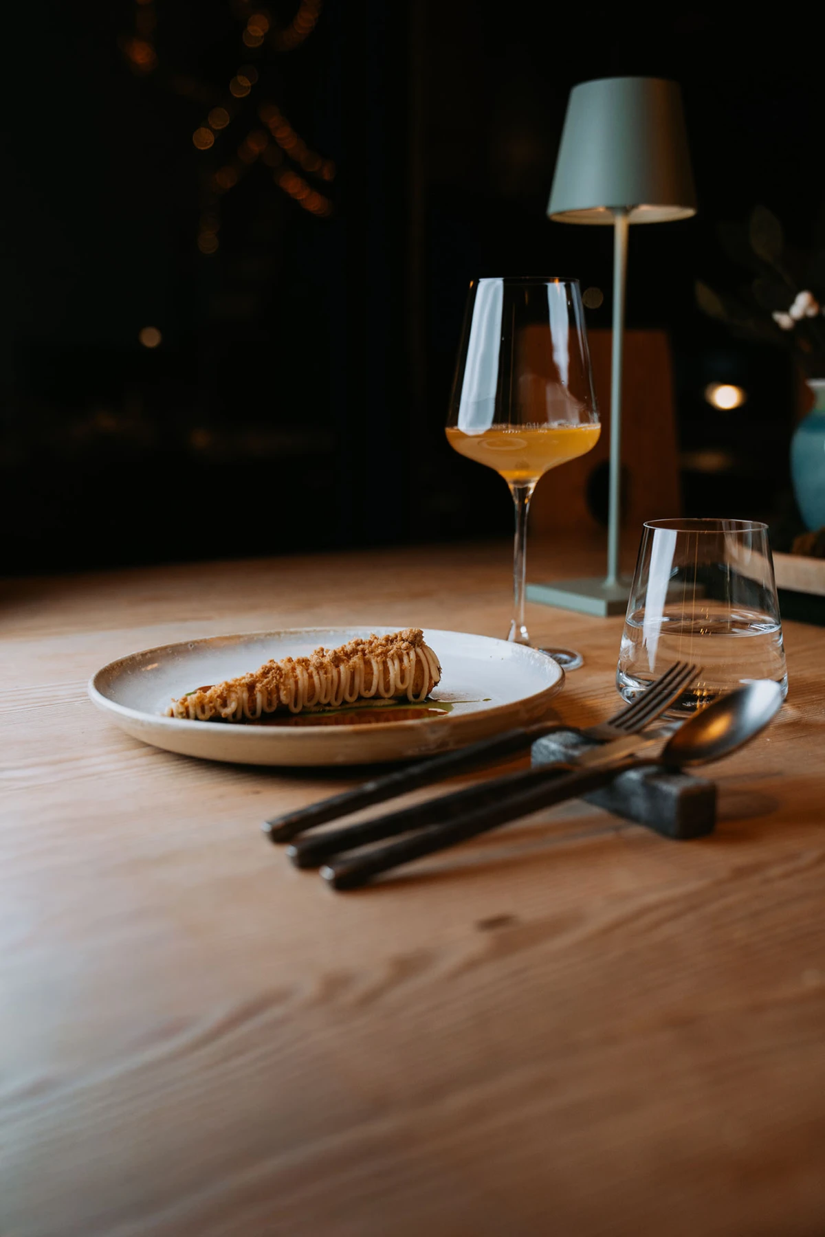 Karamellgebäckrolle liegt auf Teller, Weinglas und Wasserglas daneben, Besteck auf Holztisch, gedämpftes Restaurantlicht