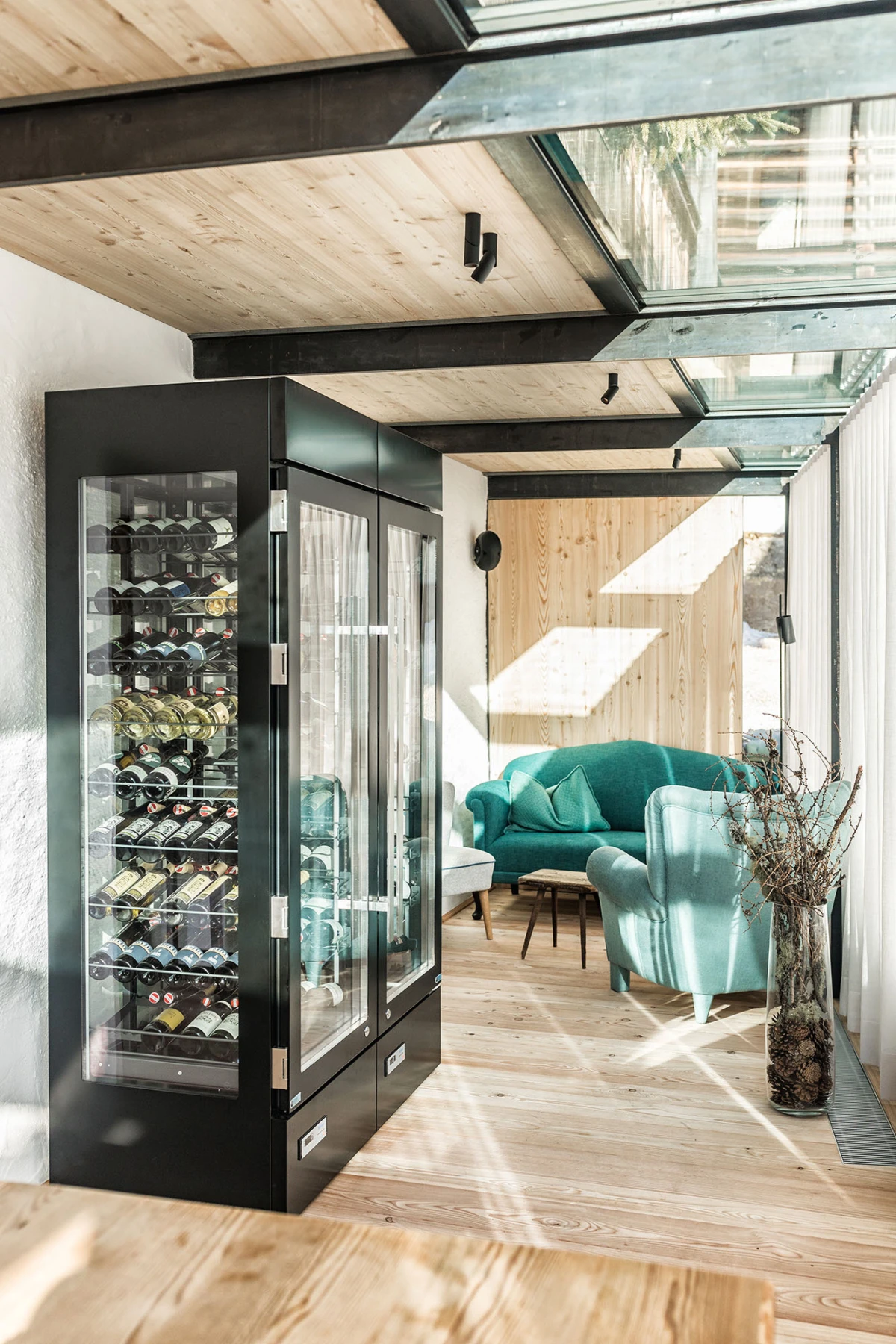 Weinkühlschrank mit Glasfront in einem lichtdurchfluteten Wintergarten; tealfarbene Sitzecke mit Sofa und Sessel auf Holzboden.