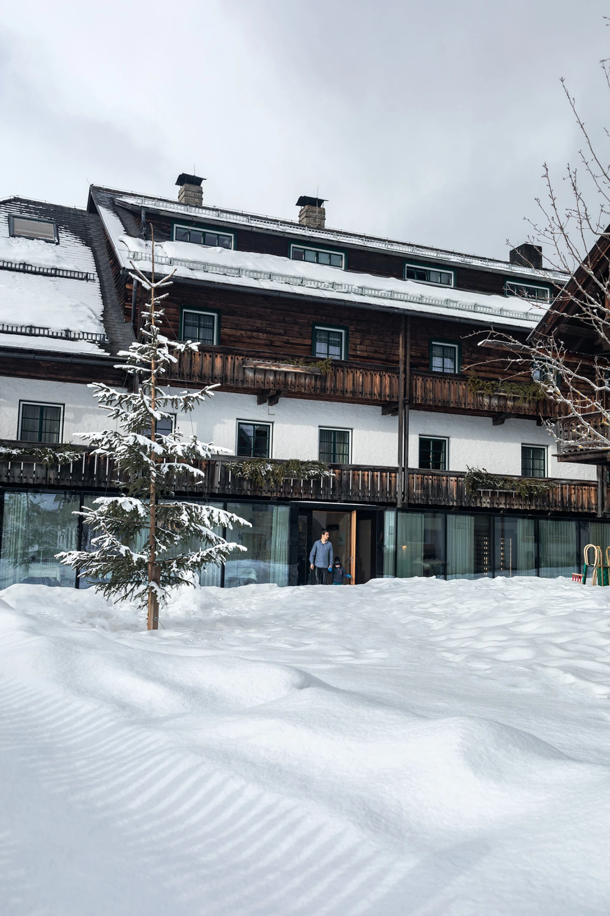 Person am Eingang eines verschneiten, mehrstöckigen Chalet-Hotels aus Holz; davor schneebedeckte Fläche und Tannenbaum.