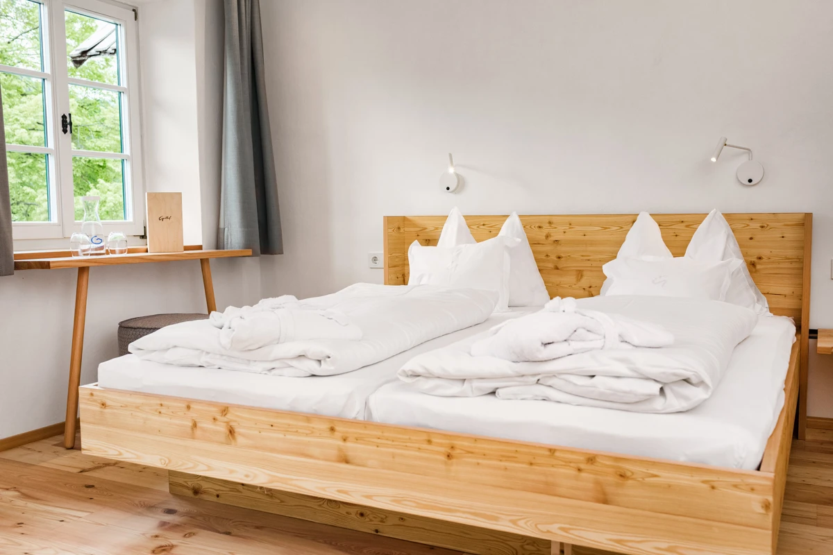 Doppelbett aus hellem Holz mit weißer Bettwäsche und gefalteten Decken; Fenster, Vorhänge und ein Schreibtisch am Fenster.