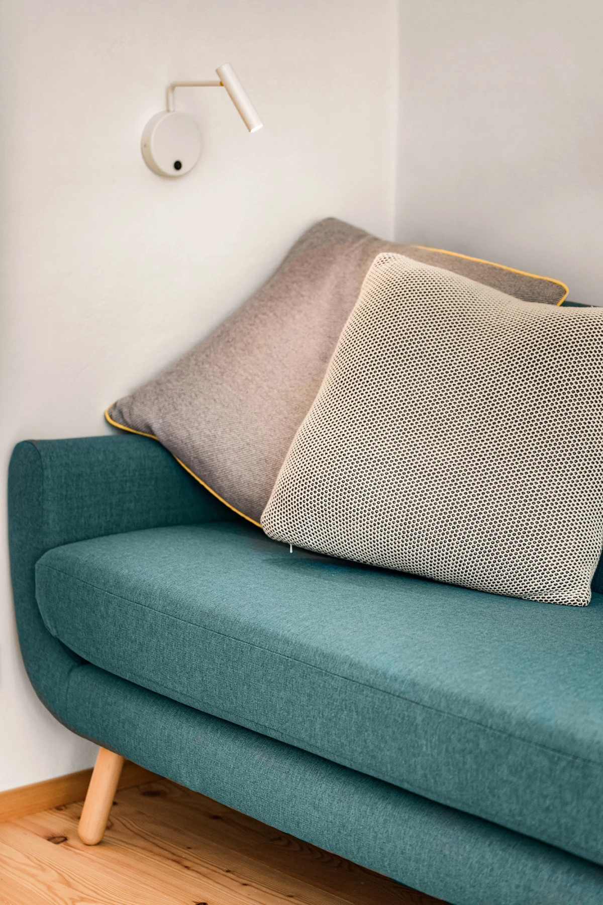 Tealfarbenes Zweisitzer-Sofa mit zwei Kissen (grau gepunktetes und hellgraues), neben Wandlampe, Holzfußboden.