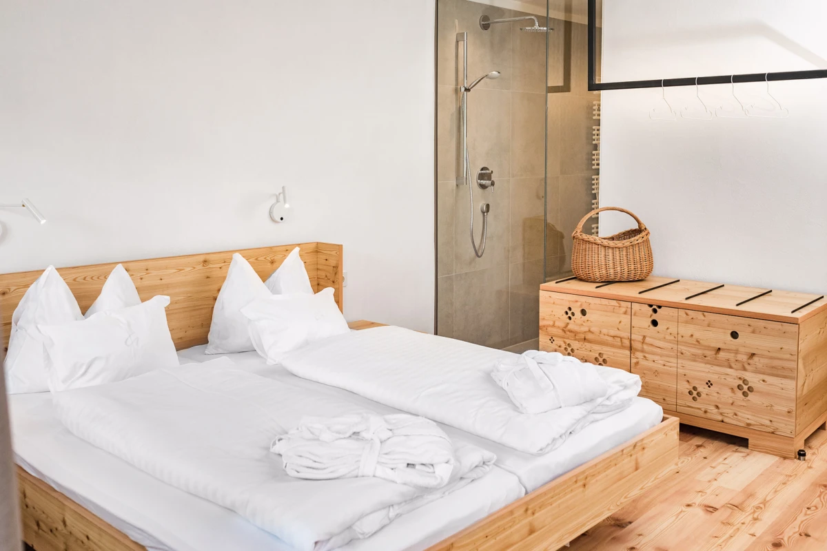 Massivholz-Doppelbett mit weißer Bettwäsche; offenes Bad mit Glasdusche, Holzkommode und Korb im hellen Schlafzimmer.