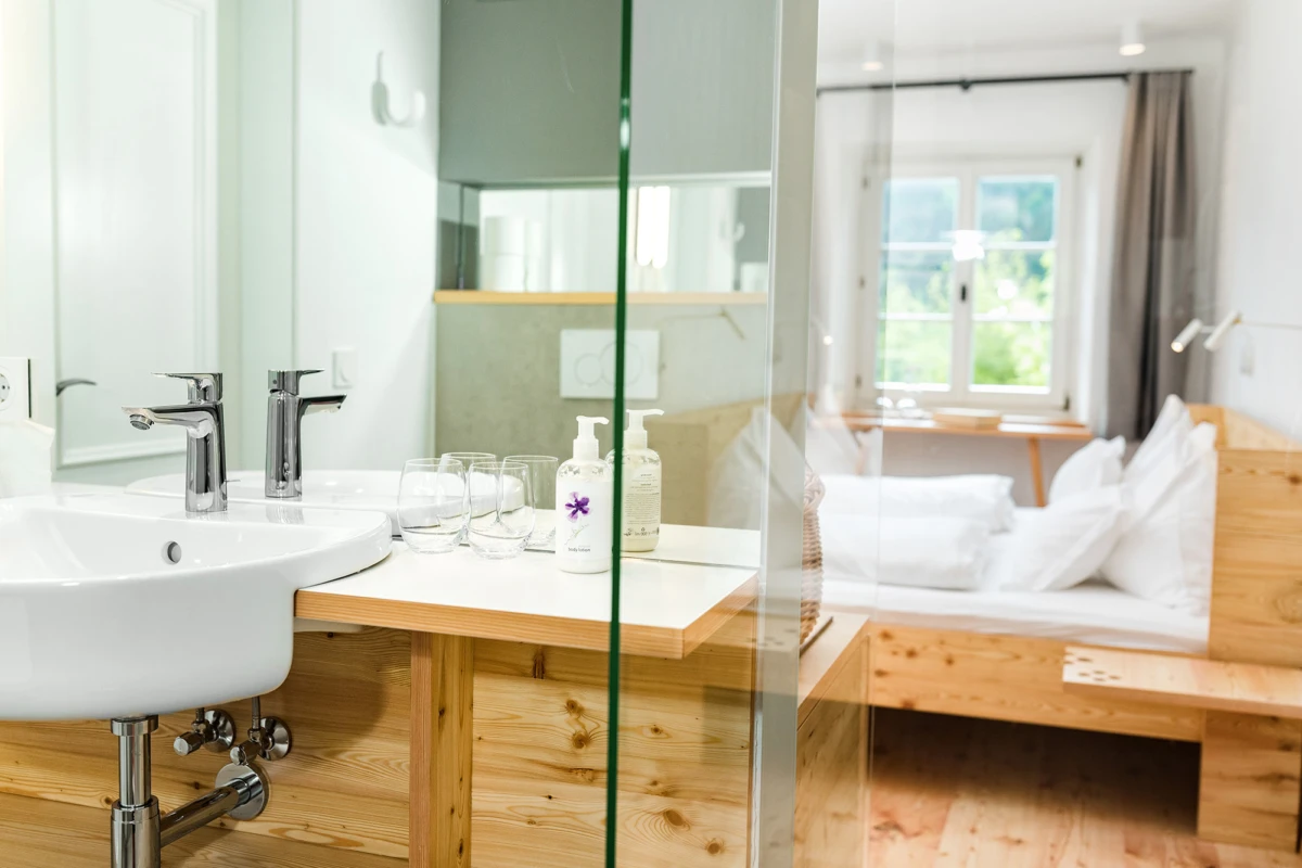 Waschbecken mit zwei Chromarmaturen auf Holzkonsole; Seifenspender und Gläser; Blick durch Glaswand auf Schlafzimmer mit Bett.
