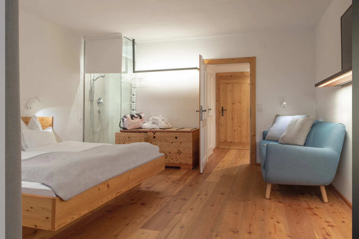 Hotelzimmer mit Doppelbett, Glasduschkabine, Holzmöbel, blauem Sofa, Holzfußboden, offene Tür zum Flur.