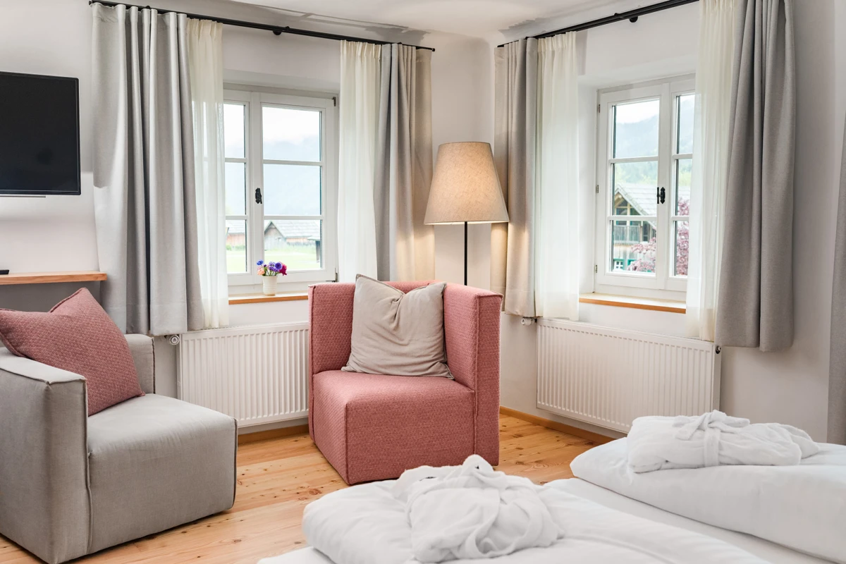 Hotelzimmer-Wohnbereich: graues Sofa, rosafarbener Sessel, Bodenlampe; zwei Fenster mit Vorhängen, Holzboden, Bademäntel auf Bett.