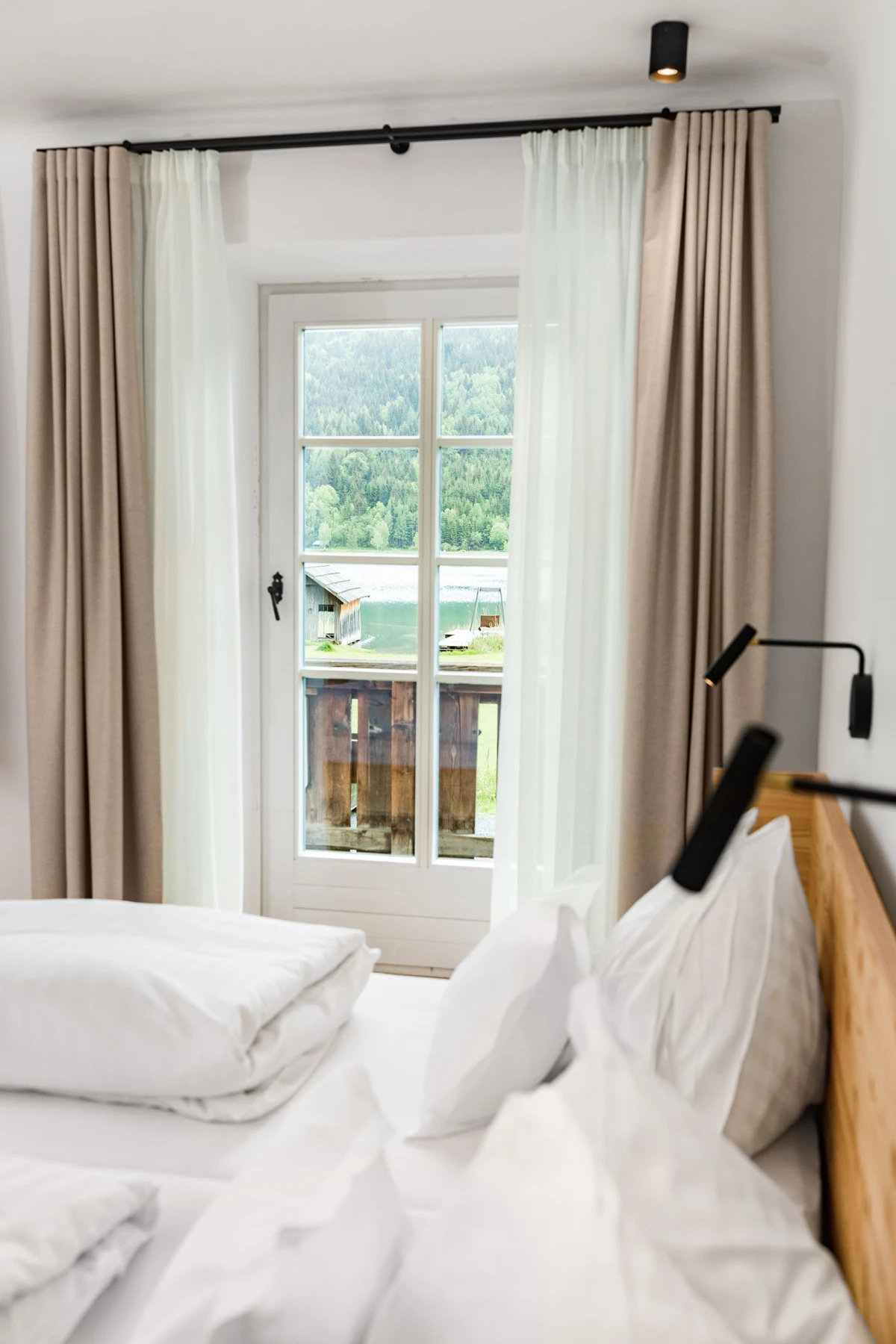 Bett mit weißer Bettwäsche und Kissen vor Balkontür; beige und weiße Vorhänge, Blick auf See und Berge.