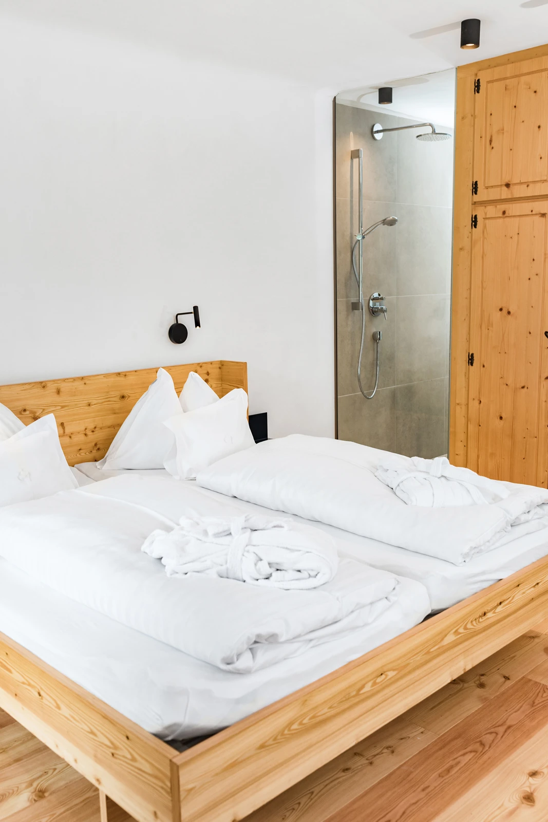 Holzbett mit weißer Bettwäsche, zwei Kissen und gefalteten Bademänteln; offener Duschbereich und Holzschrank im Hintergrund.