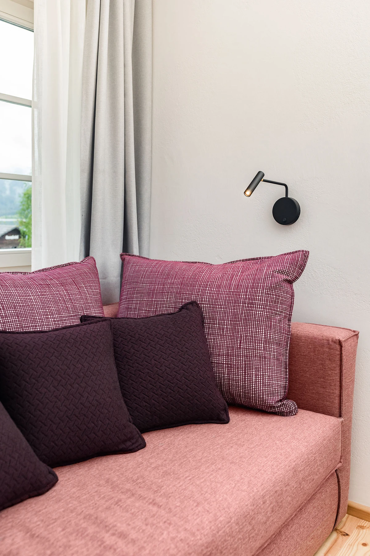 Rosa Stoffsofa mit dunkellila und karierten rosa Kissen vor Fenster mit Vorhängen; Wandlampe daneben.