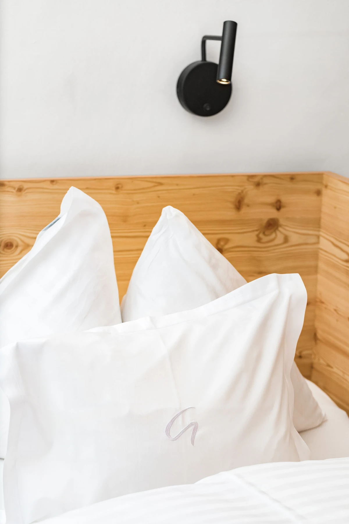 Bett mit weißen Kissen vor Holz-Kopfteil; schwarze Wandlampe darüber.