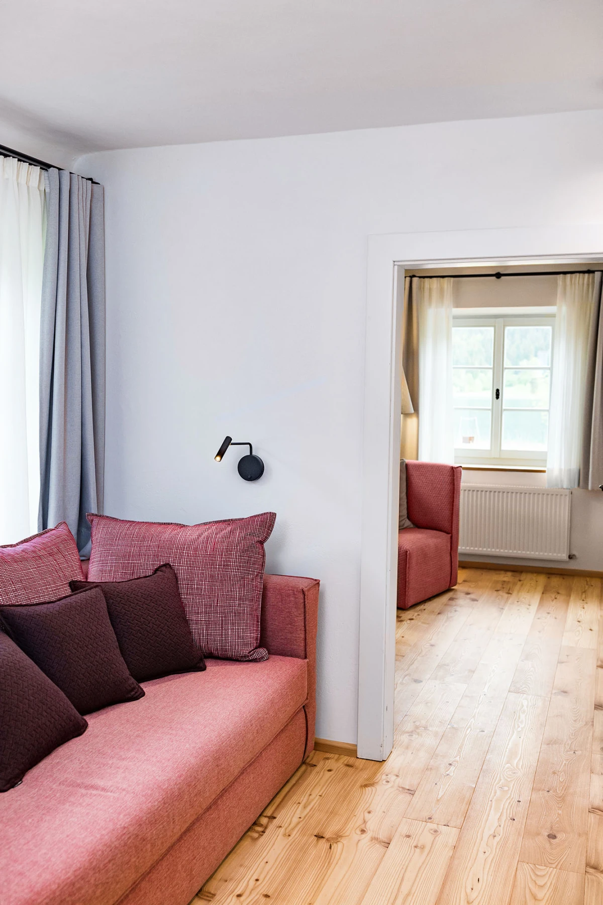Rosa Stoffsofa mit dunklen Kissen im Wohnzimmer; schwarze Wandlampe, helle Vorhänge. Durchgang zu einem zweiten Raum mit rotem Sessel und Fenster.
