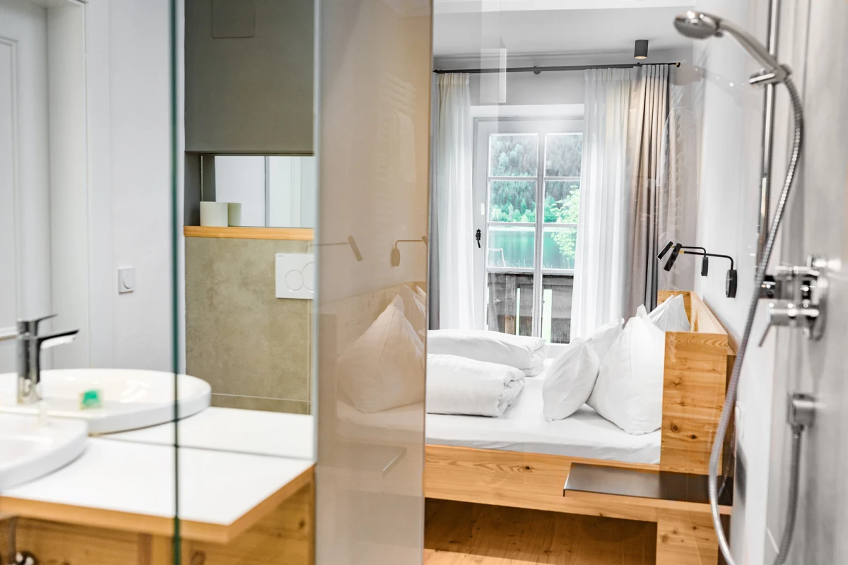 Modernes Schlafzimmer mit Holzbett und weißer Bettwäsche, Glasabtrennung zum Bad; Waschbecken links, Fenster mit Vorhängen.