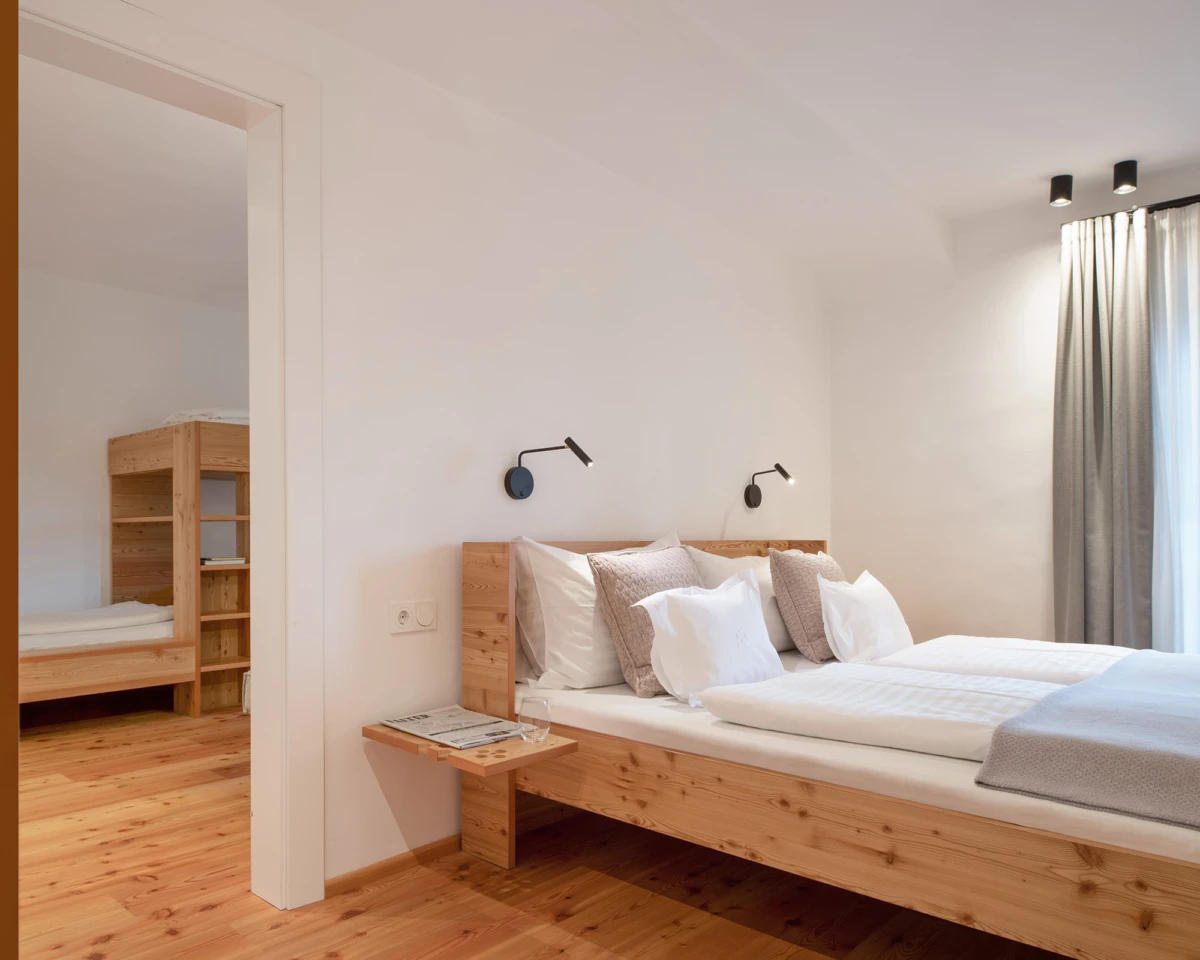 Holz-Doppelbett mit weißer Bettwäsche, grauen Kissen, Wandlampen, Holzfußboden, graue Vorhänge; Durchgang zu zweitem Zimmer mit Etagenbett.