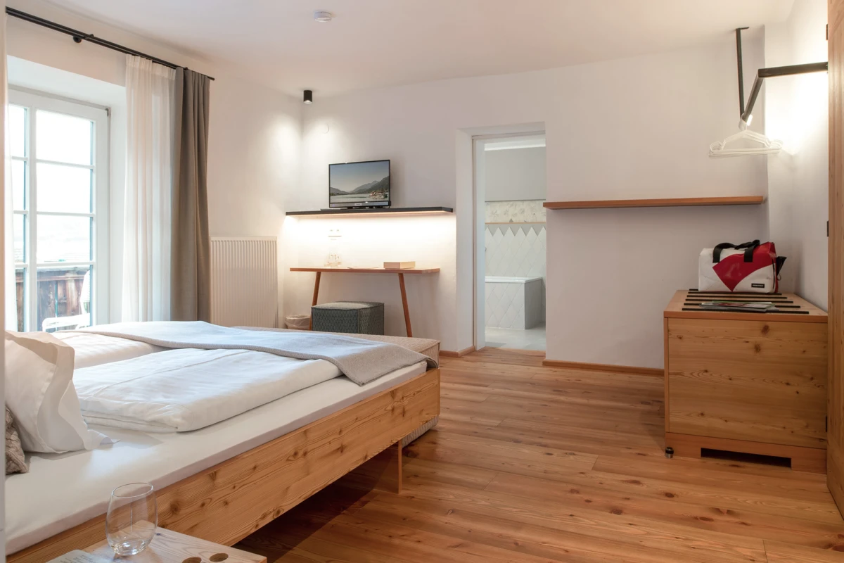 Holzfußboden-Schlafzimmer mit Doppelbett, weißer Bettwäsche; links Balkonfenster, rechts Kommode mit rotem Rucksack, Schreibtisch mit Fernseher.