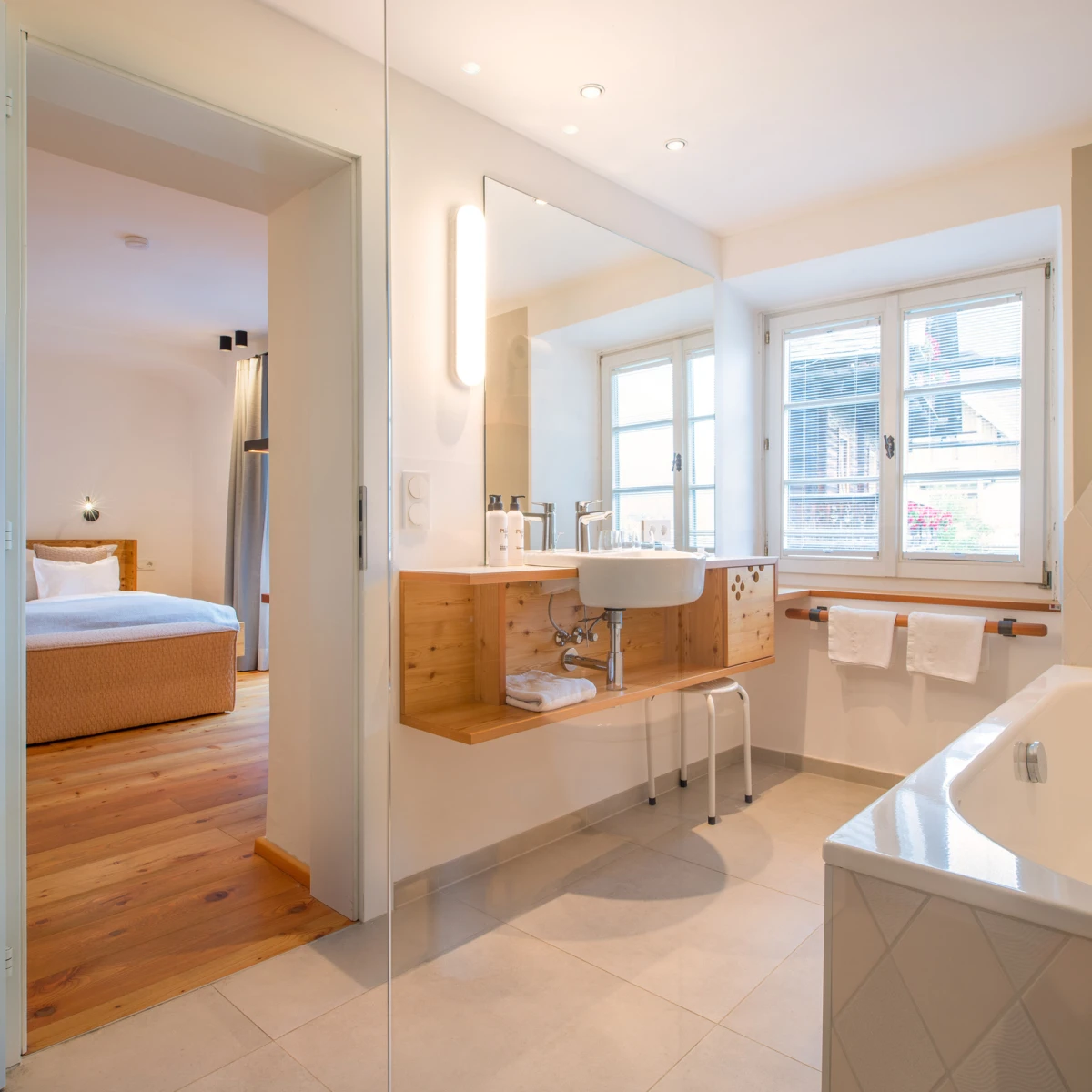 Helles Badezimmer mit Waschbecken auf Holz-Waschtisch, Spiegel, Badewanne und Fenster; angrenzendes Schlafzimmer im Türdurchgang sichtbar.