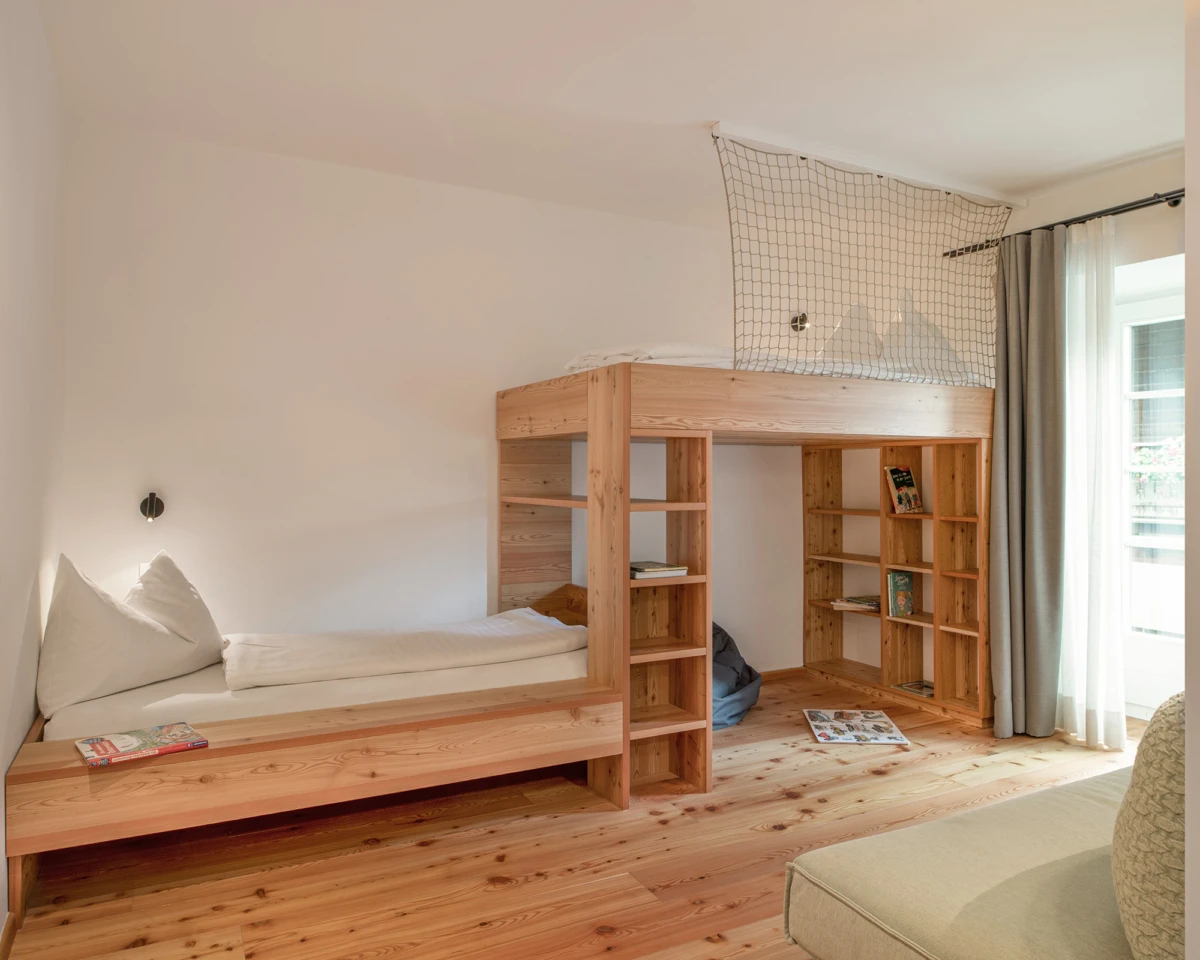 Holz-Loftbett mit eingebauten Regalen und Netzschutz am oberen Bett; darunter Liegefläche, Bücherregale, großes Fenster mit Vorhängen.