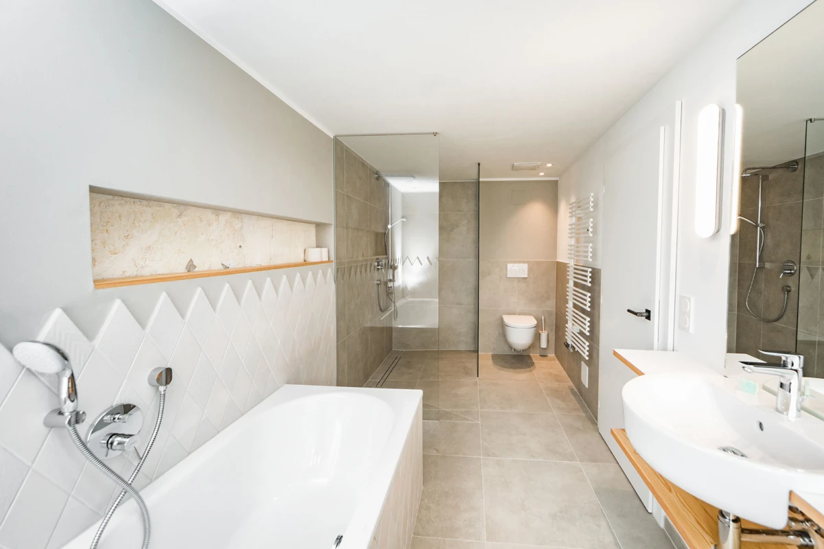 Modernes Badezimmer: linke Badewanne, mittlere Dusche, hintere Toilette, rechtes Waschbecken mit Spiegel, Boden- und Wandfliesen.