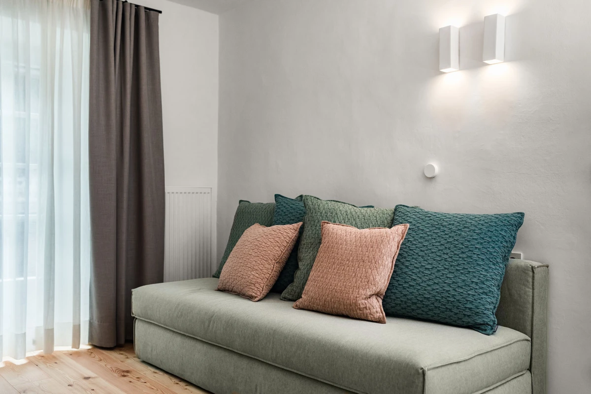 Grünes Sofa mit pfirsichfarbenen und türkisfarbenen Kissen steht vor heller Wand; Vorhänge links, moderne Wandlampen oben.