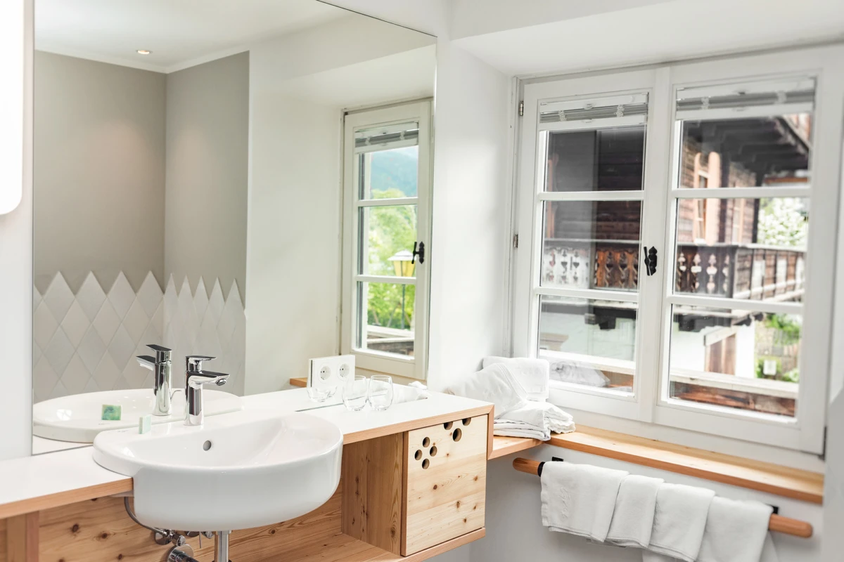 Waschbecken mit Chromarmaturen auf Holzkonsole, Spiegel darüber; Fensterbank mit Handtüchern, Blick auf Außenbalkon.