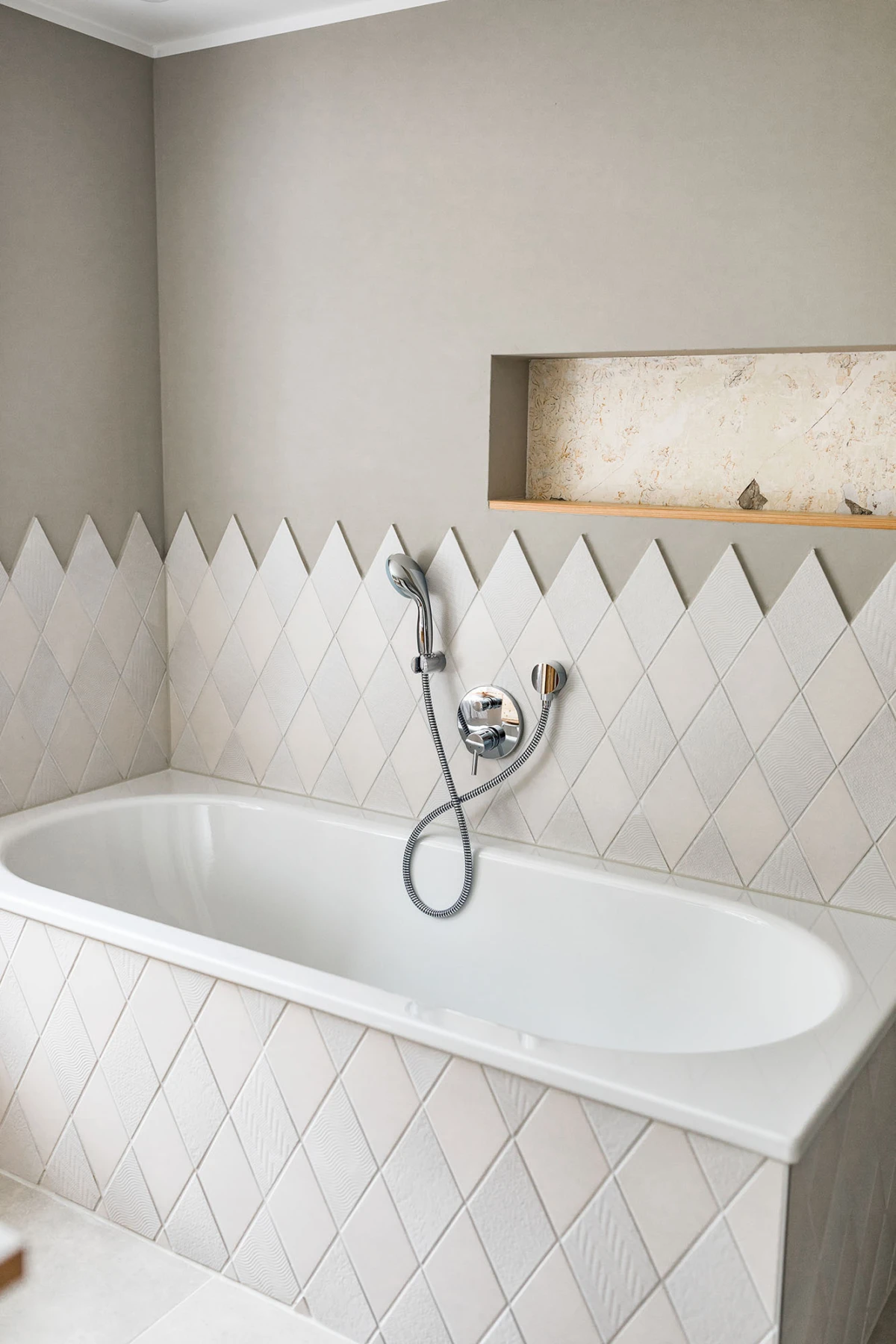 Weiße Badewanne, beige Wand, Fliesen mit Diamantmuster, Handbrause mit Schlauch an Armatur, Wandnische mit Steindekor.