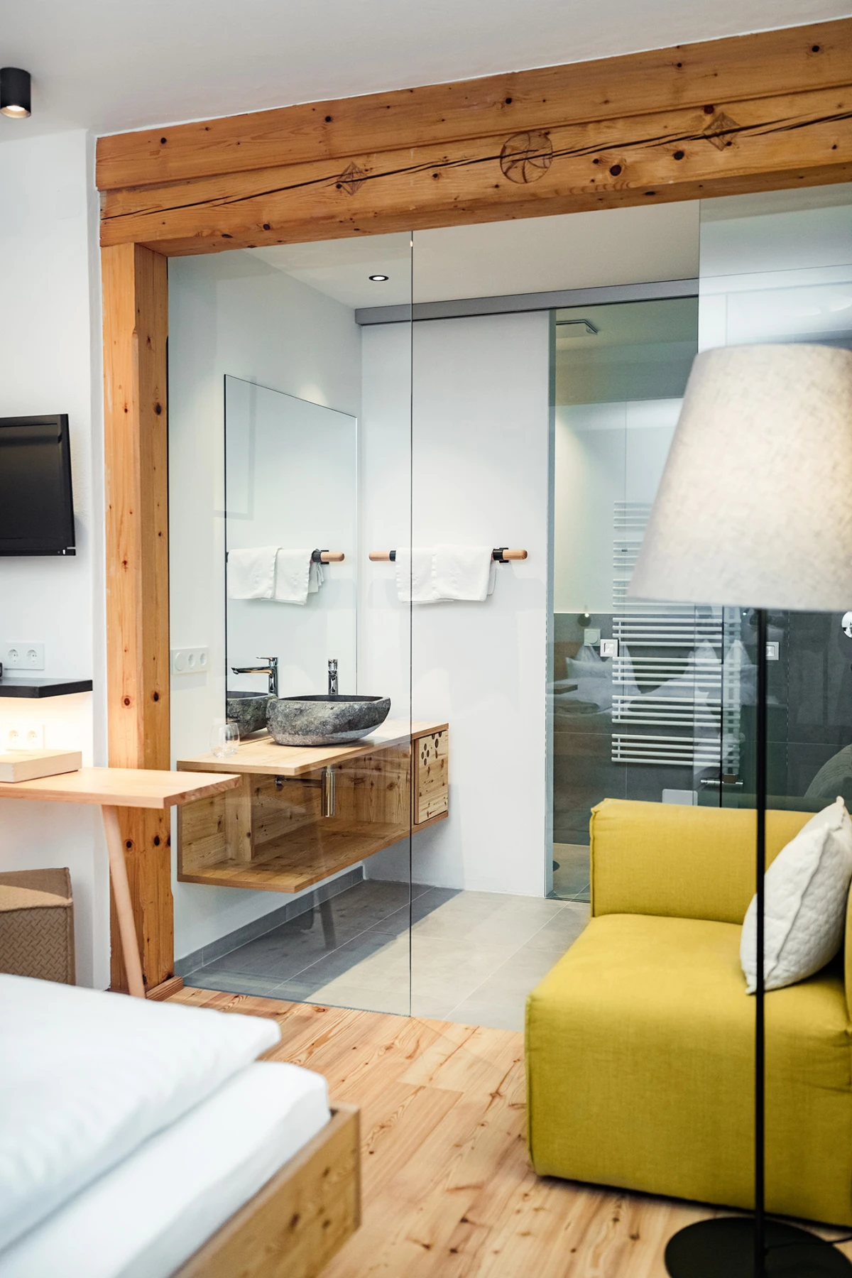 Offenes Hotelzimmer mit Glas-Trennwand zum Bad; Stein-Waschbecken auf Holz-Waschtisch, weiße Handtücher, gelbes Sofa, Holzboden und Holzbalken.