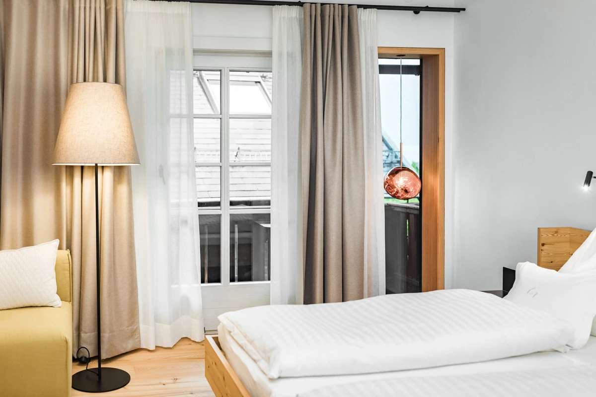 Doppelbett mit weißer Bettwäsche, Holz-Kopfteil, Stehlampe, beige und weiße Vorhänge, Balkontür mit kupfernem Pendellicht.