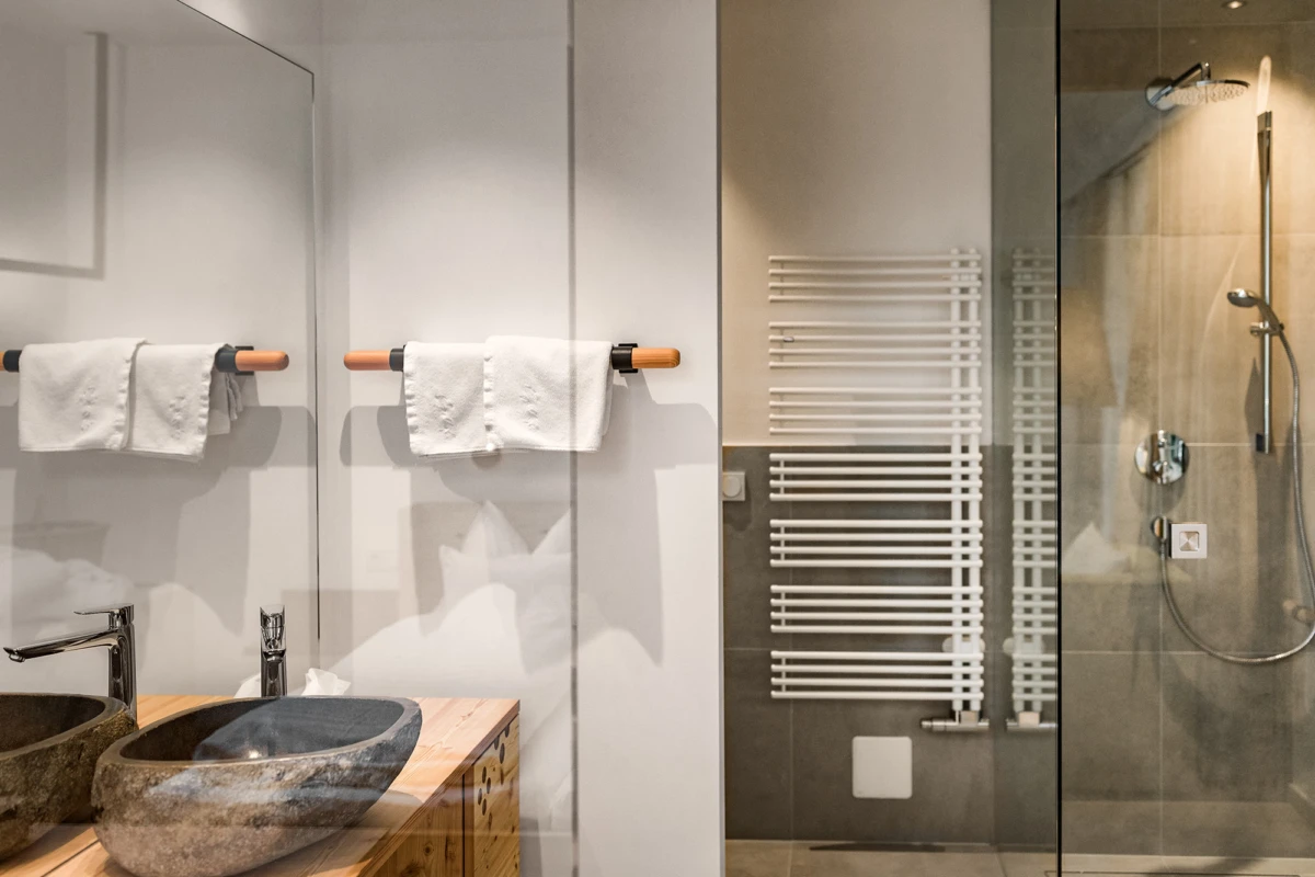 Zwei Steinwaschbecken auf einer Holzplatte, weiße Handtücher an einer Holzstange; Glasdusche mit Handbrause; horizontaler Handtuchheizkörper an der Wand.