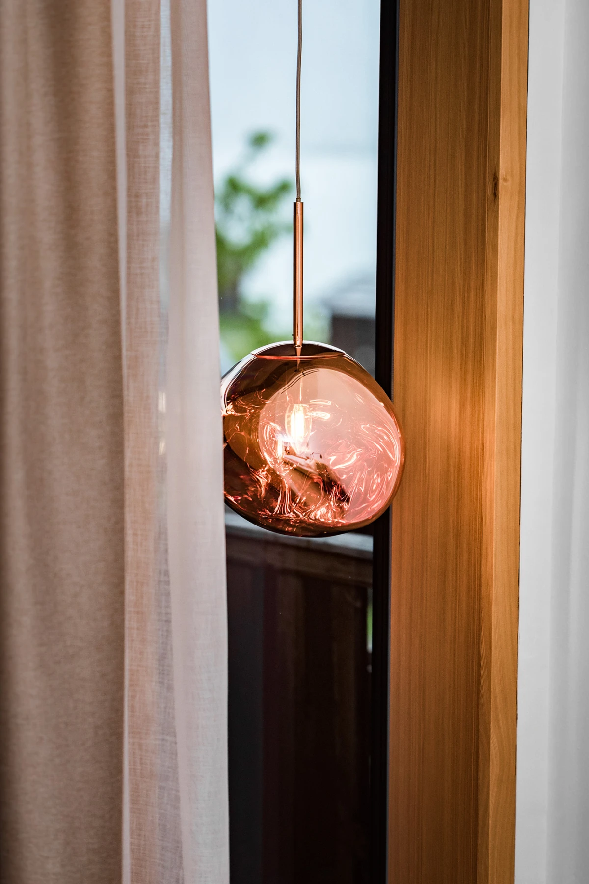 Kupferfarbene Glaskugel-Pendelleuchte hängt an Kabel vor Fensterrahmen mit Vorhang.