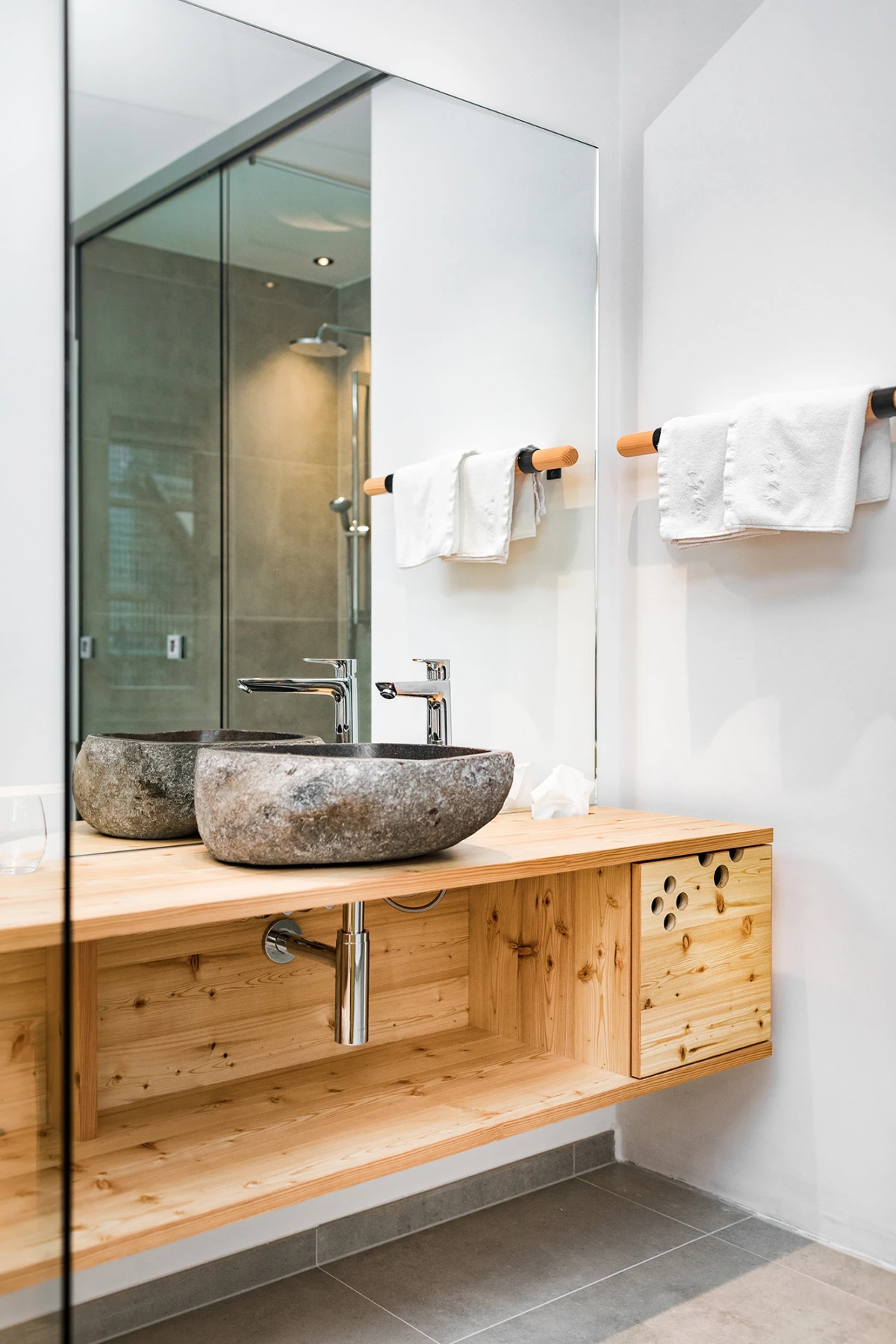 Zwei Stein-Waschbecken auf einer Holzplatte in modernem Badezimmer, Spiegel, Handtücher an Halter, Glasdusche im Hintergrund.