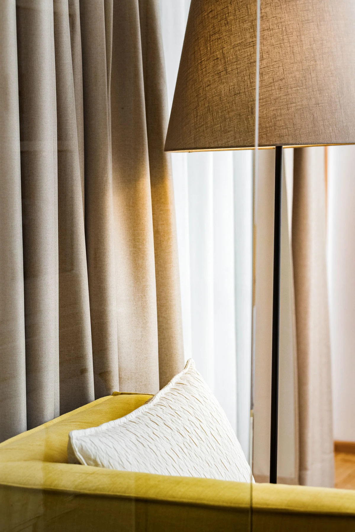 Senfgelbes Sofa mit weißem Kissen, neben braunem Bodenlampenschirm; beige Vorhänge im Hintergrund