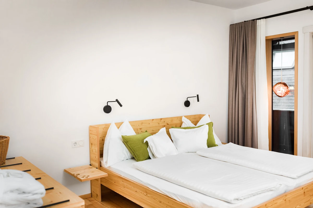 Holz-Doppelbett mit weißer Bettwäsche und grünen Kissen, zwei schwarze Wand-Leseleuchten, seitlicher Nachttisch, großes Fenster mit Vorhängen
