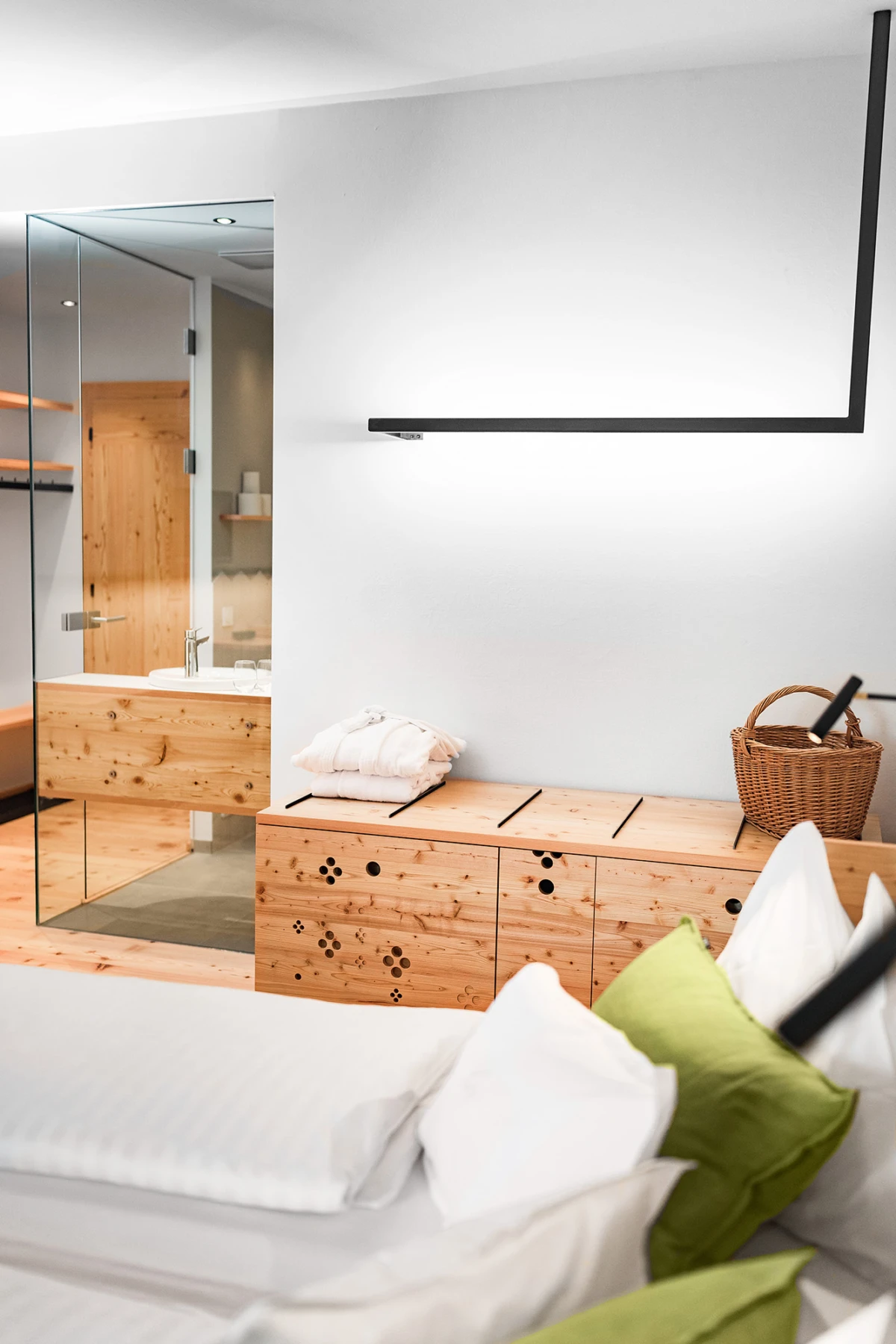 Schlafzimmer mit weißem Bett und grünen Kissen, Holzkonsole mit Korb; Blick auf Badezimmer hinter Glaswand.