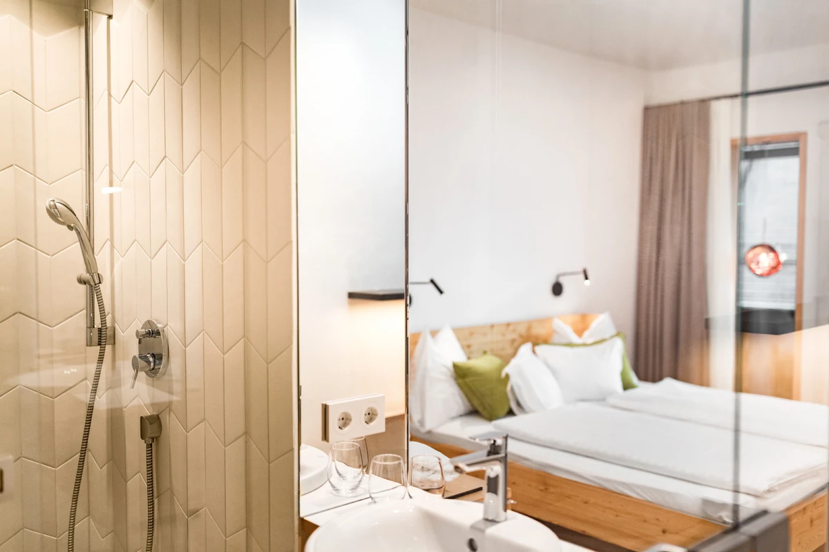 Hotelzimmer mit offener Dusche, Glaswand zum Schlafzimmer; Bett mit weißen Laken und grünen Kissen.