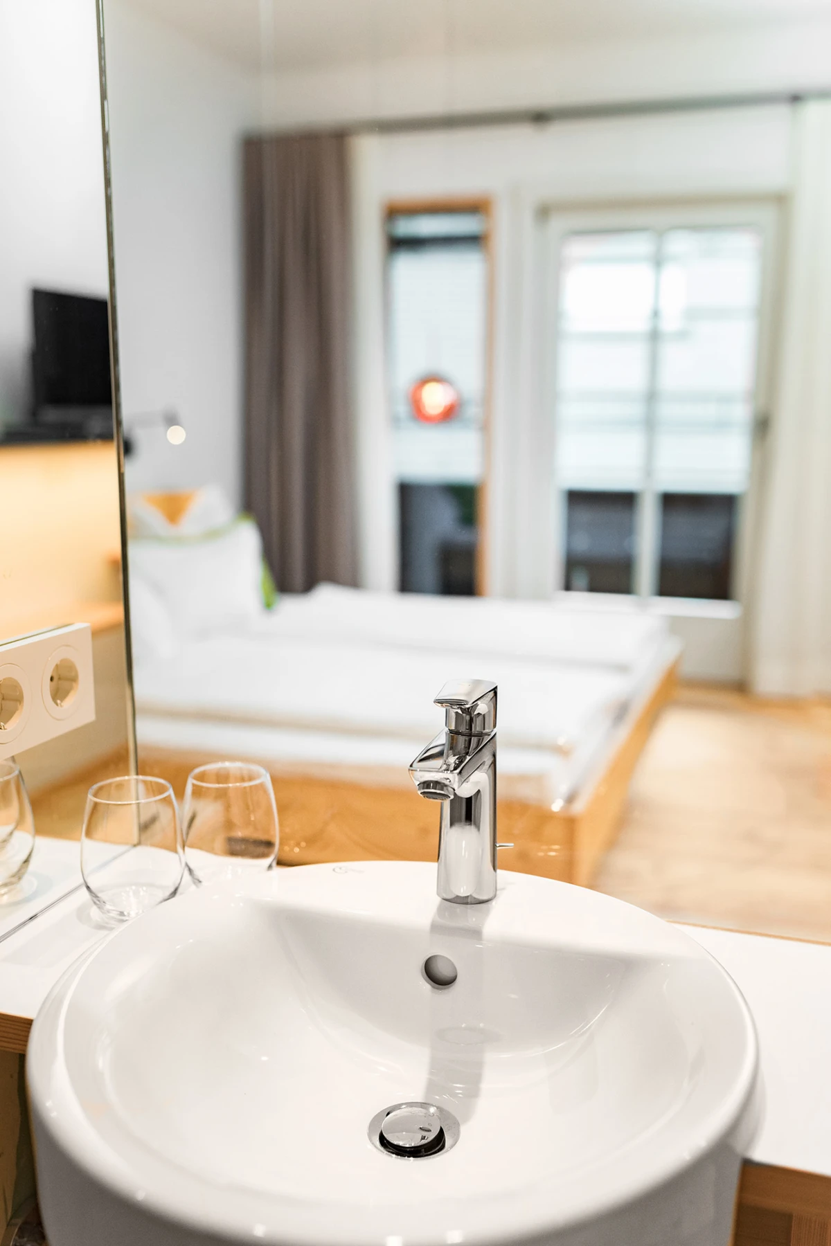 Rundes weißes Waschbecken mit glänzender Chromarmatur; zwei Weingläser daneben; Hintergrund: Hotelzimmer mit Bett, Vorhängen und Fenster.