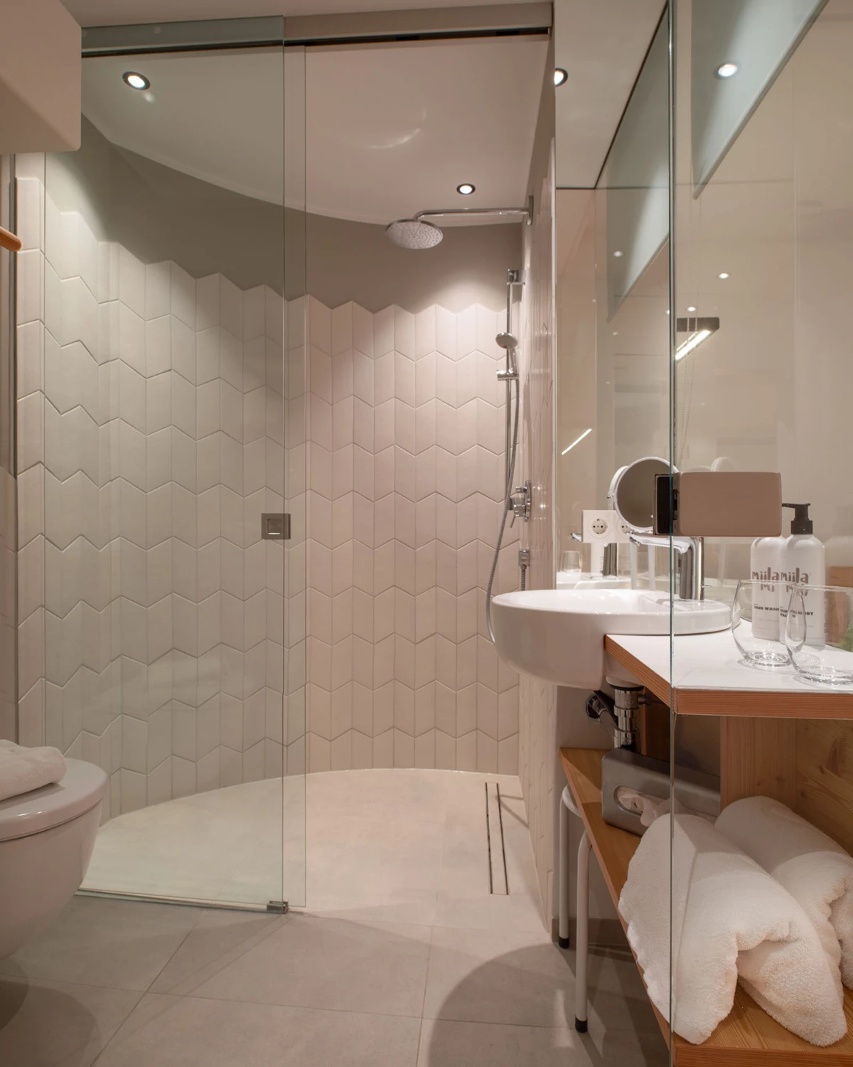 Modernes Badezimmer mit begehbarer Glasdusche, sechseckigen Wandfliesen, Waschbecken, Spiegel und Holzablage mit Handtüchern.