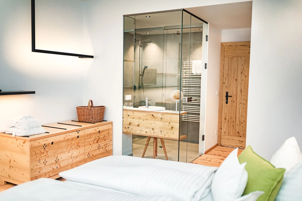 Schlafzimmer mit Holzkommode, Wäschekorb und weißer Bettwäsche; Glaskabine des Badezimmers mit Dusche rechts sichtbar.
